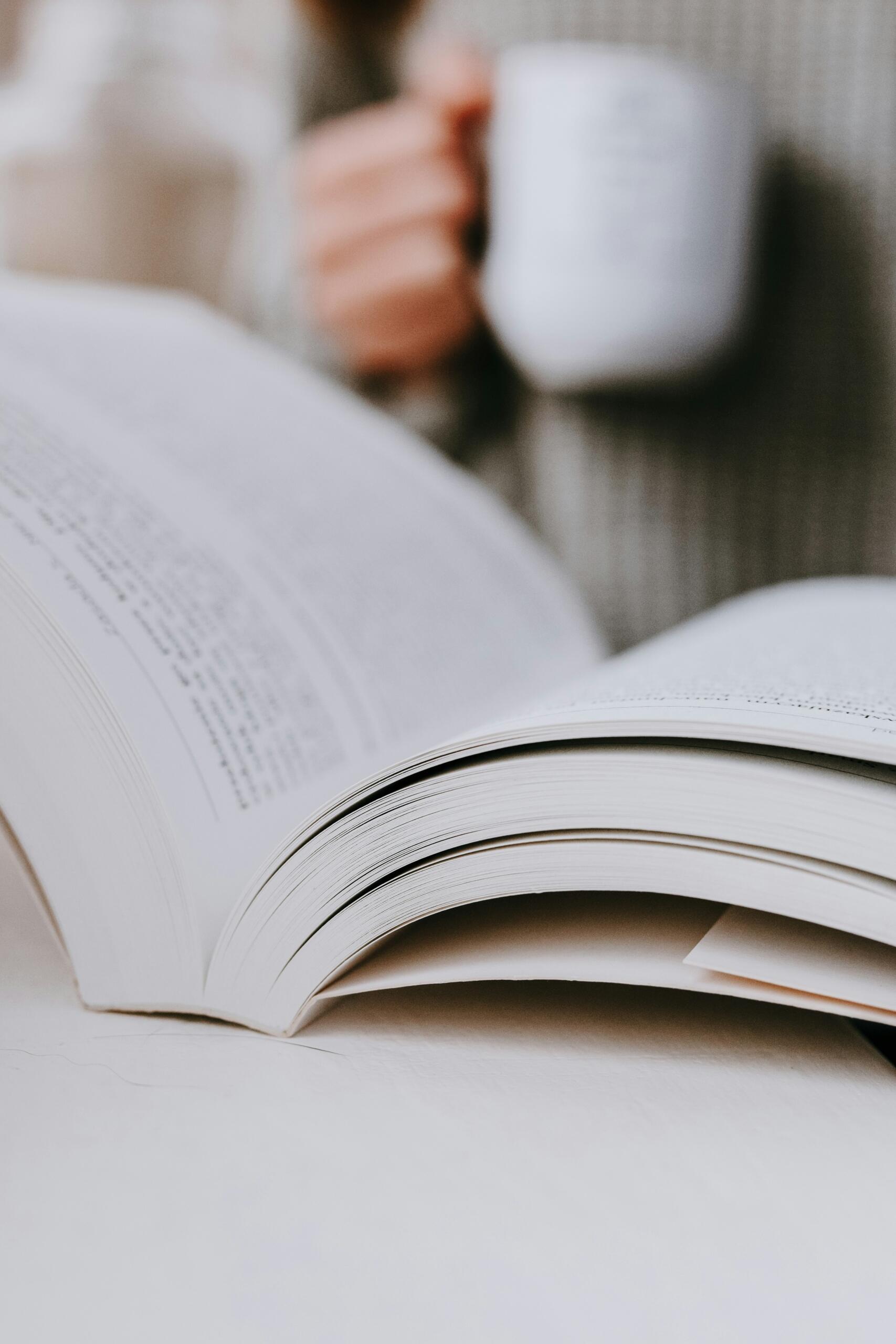 Atvērta grāmata uz galda, kamēr fonā redzama roka ar kafijas krūzi.