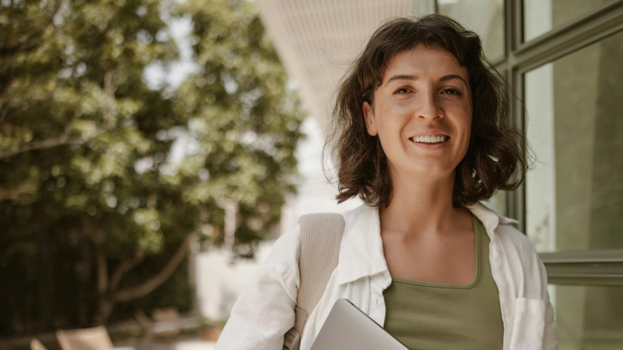 sieviete baltā virsblūzē tur rokās portatīvo datoru un smaida