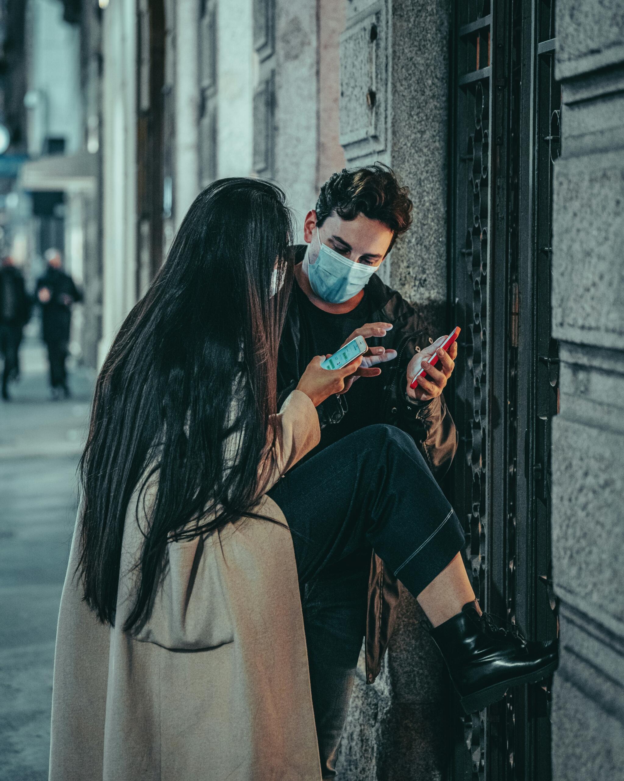 Divi cilvēki pie ēkas sienas skatās telefonos un sarunājas.