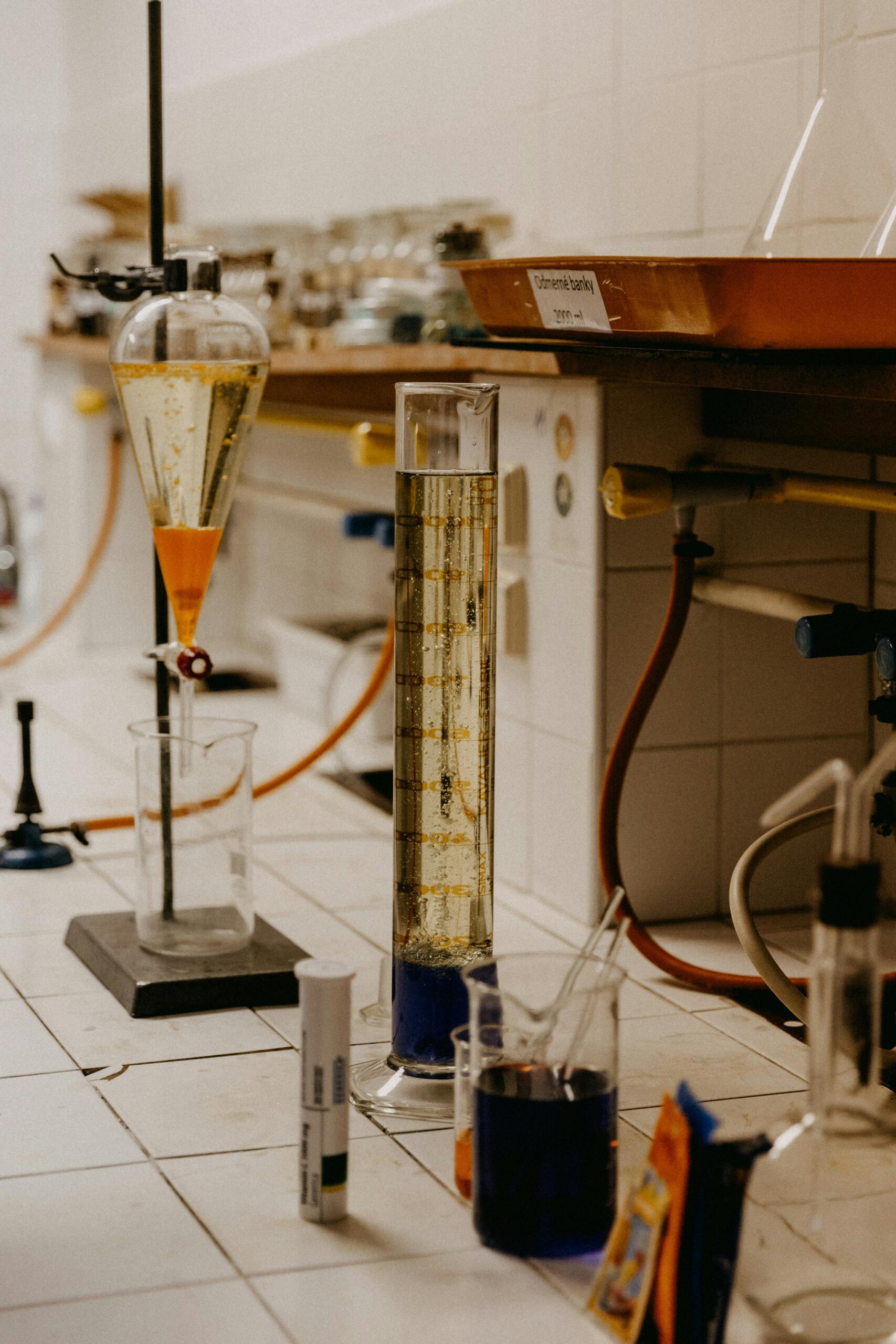 Uz galda izkārtoti dažādi laboratorijas trauki ar krāsainiem šķidrumiem pētījumiem.
