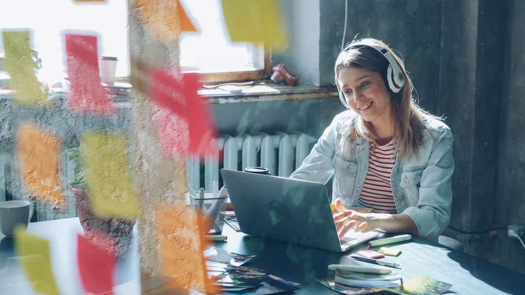 Mlada kreativna poslovna žena sa slušalicama na ušima sluša muziku i pleše dok radi za laptopom u modernom kancelarijskom prostoru. U prvom planu nalazi se čaša sa šarenim naljepnicama.