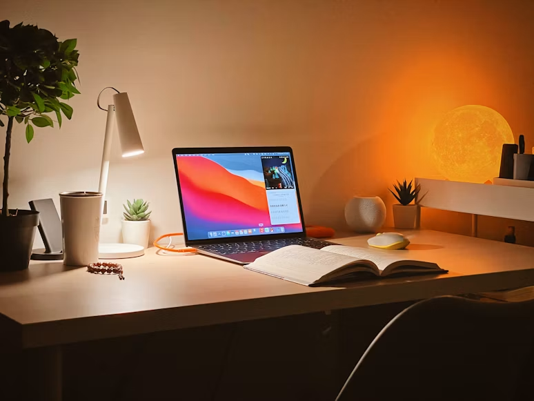 MacBook Pro nalazi se na bijelom drvenom stolu u prijatnom i mirnom ambijentu. Jednostavan prizor stvara osjećaj tišine, opuštenosti i spokojne noći.