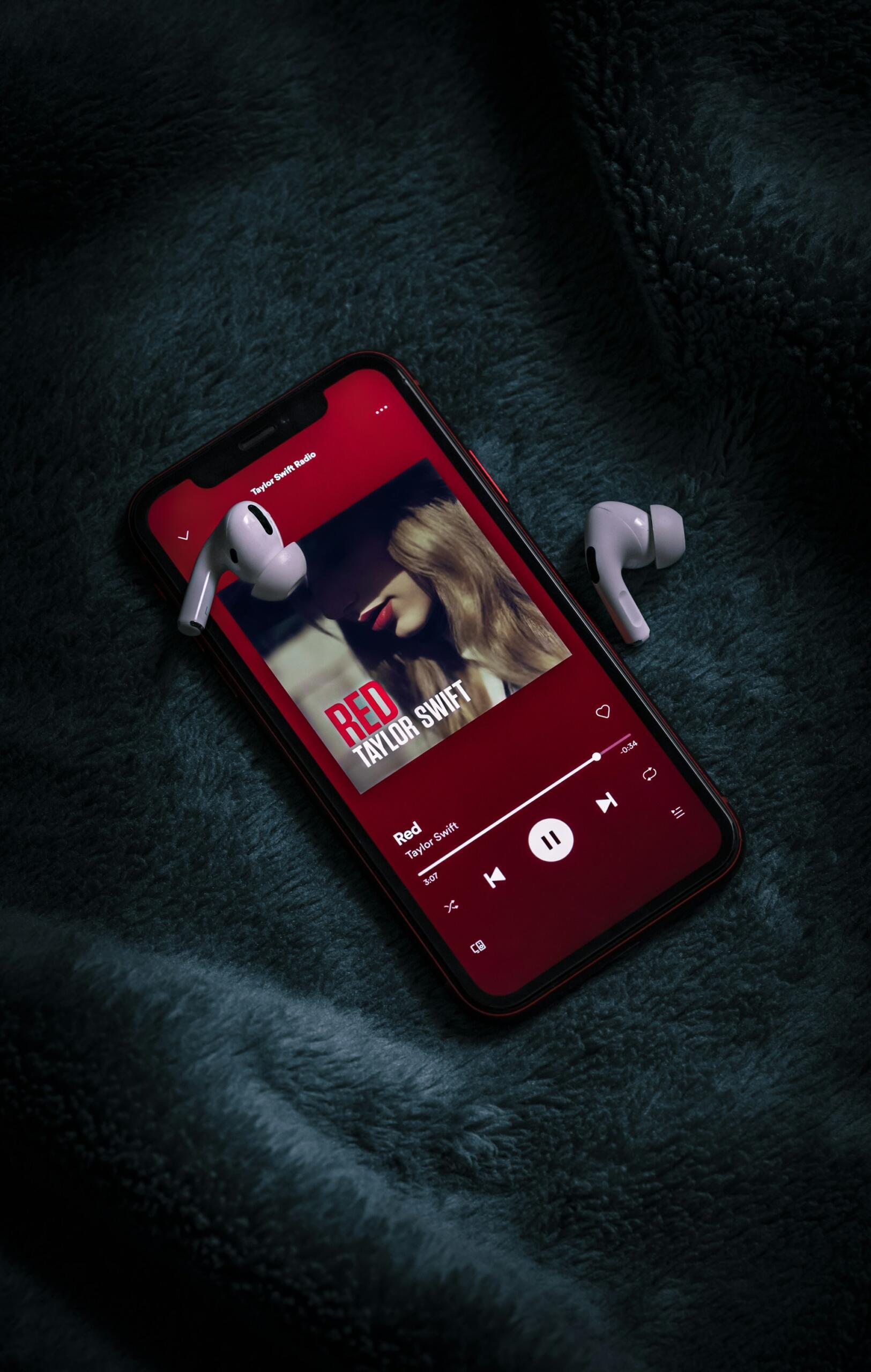 Pametni telefon na kojem je prikazana naslovna omota albuma „Red“ Taylor Swift, sa AirPods slušalicama pored njega, položen na mekom, tamnoplavom ćebetu.
