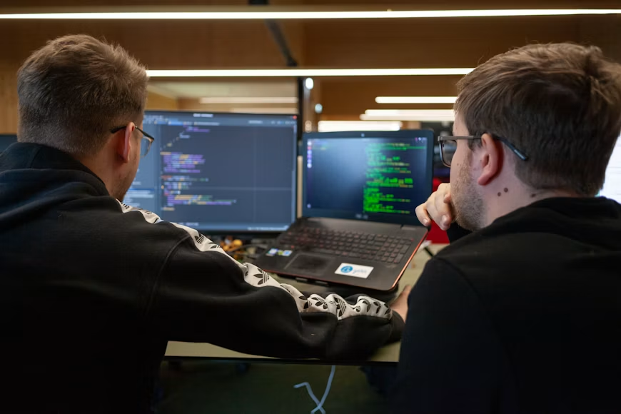 Na slici su prikazani IT stručnjaci koji pažljivo posmatraju i analiziraju linije programskog koda otvorene na ekranu laptopa i dodatnog monitora. Scena dočarava dinamičnu svakodnevicu i saradnju u svetu programiranja, gde programeri rešavaju tehničke izazove, testiraju funkcije i razvijaju savremena softverska rešenja u modernom radnom okruženju.