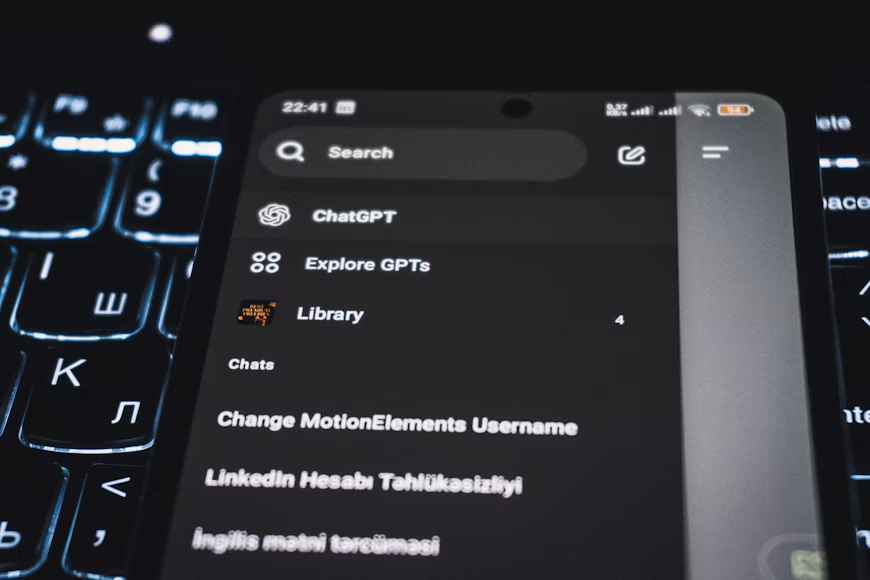 Fotografija iz blizine prikazuje ekran pametnog telefona položen na tastaturu računara. Na ekranu telefona jasno je vidljiv interfejs aplikacije ChatGPT, dok se u pozadini naziru tasteri i moderan radni prostor.