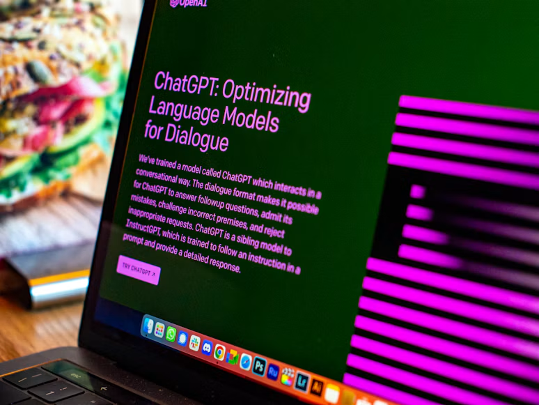 Fotografija autora Emiliana Vittoriosija prikazuje moderan MacBook računar otvoren na drvenom radnom stolu. Na ekranu je prikazana početna stranica ChatGPT-a, popularnog AI jezičkog modela namenjenog komunikaciji, olakšavanju pisanja i pružanju informacija.