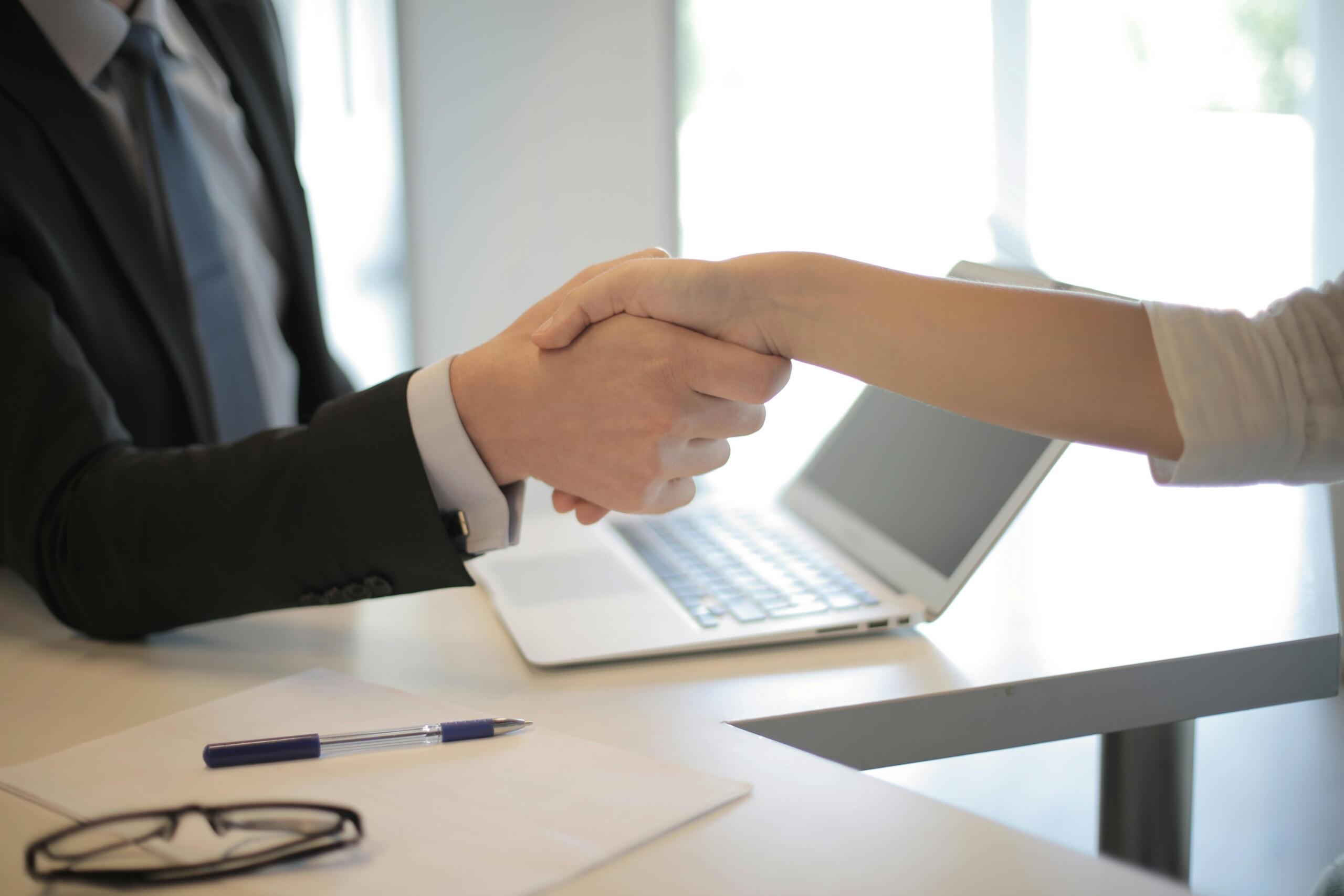 Close‑up of two people shaking hands across a desk with a laptop; one wears a black suit, the other a light top, and a pen, paper and glasses rest on the table.