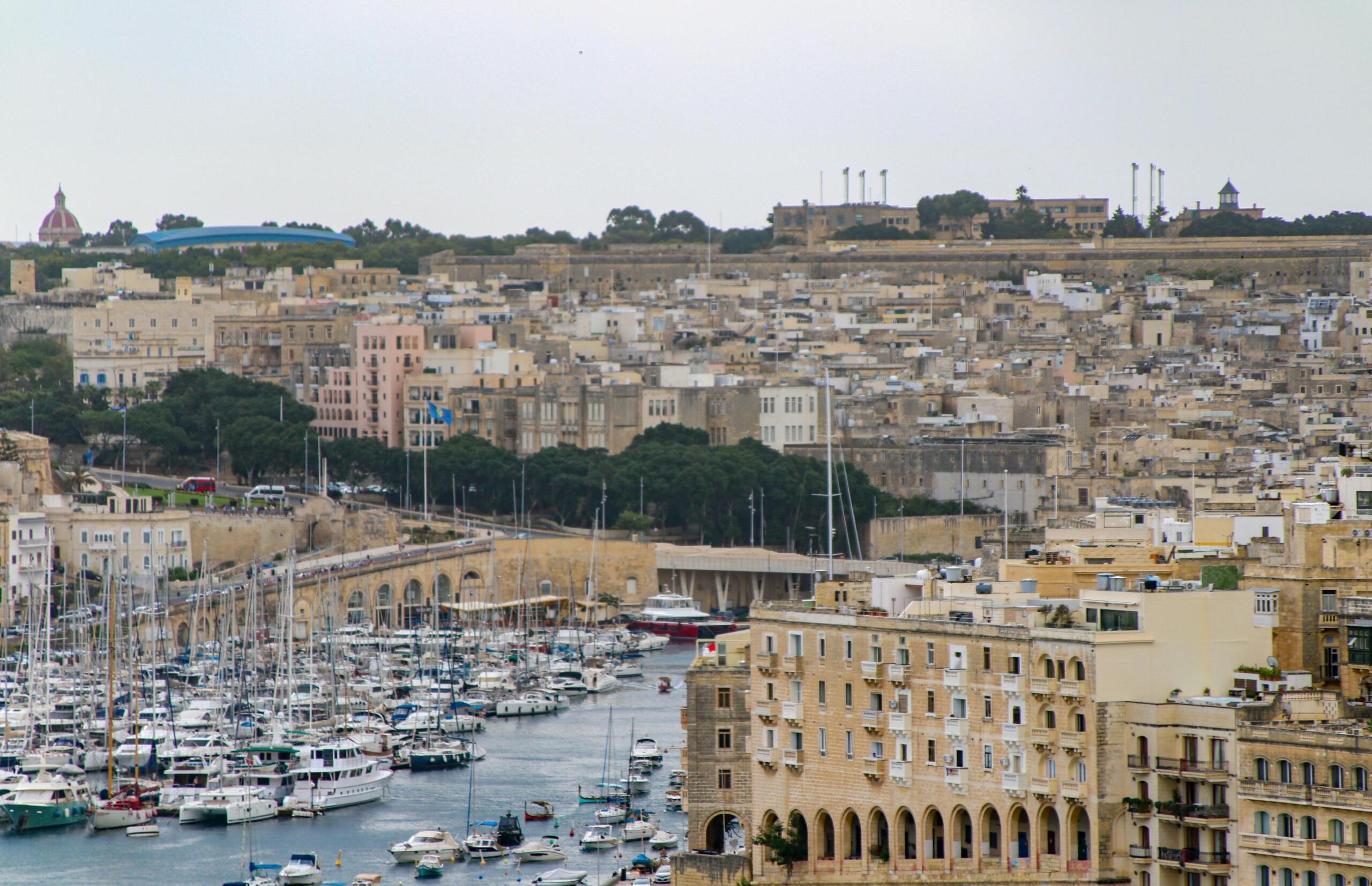A harbour packed with white yachts and sailboats beneath a hillside of tightly clustered stone and pastel buildings; a bridge spans the inlet and a domed building and a stadium roof rise on the hill.