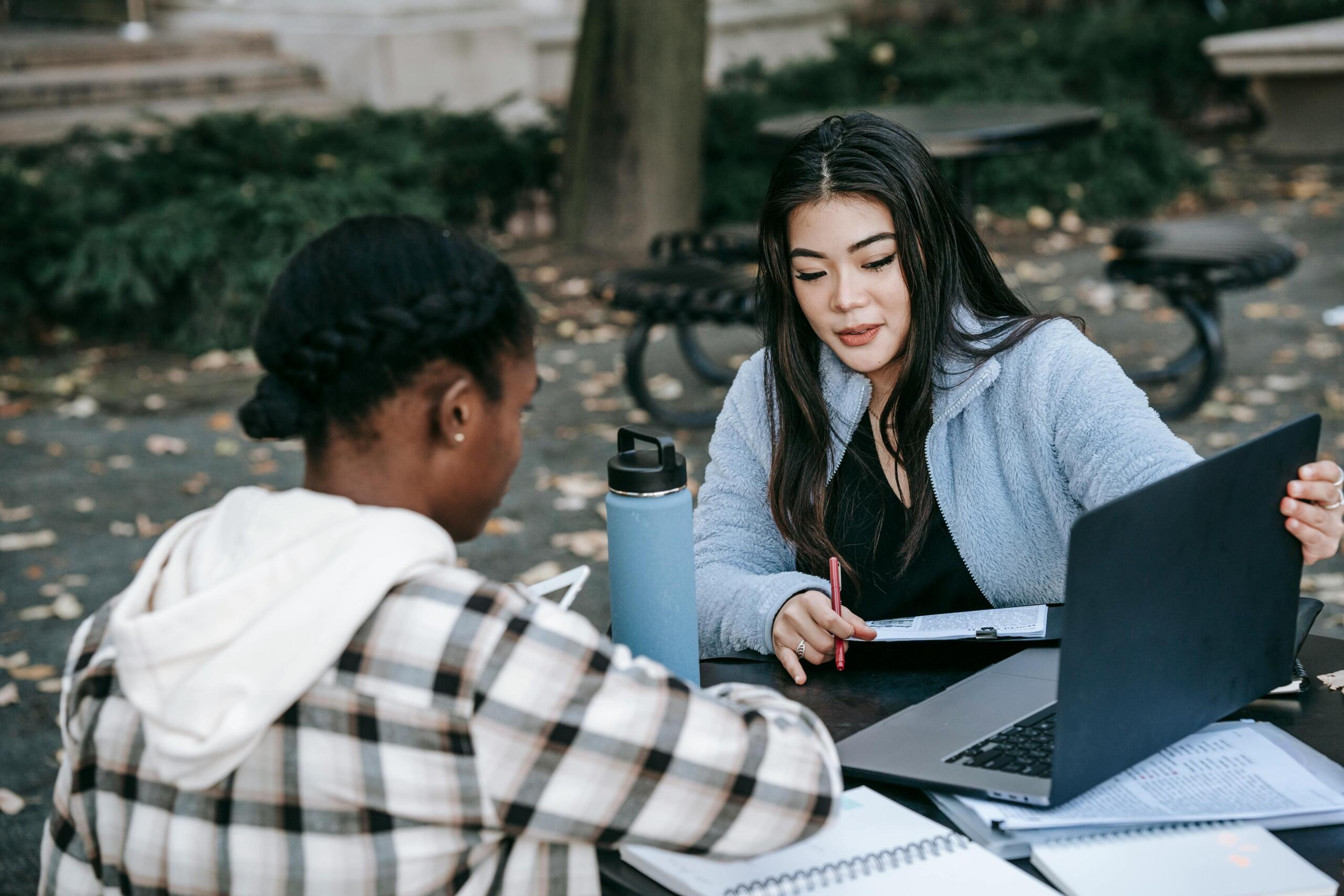 Two female students sit at an outdoor table with notebooks, a laptop and a blue water bottle; one faces away in a plaid jacket while the other, in a light blue fleece, holds a notebook and pen amid fallen leaves.