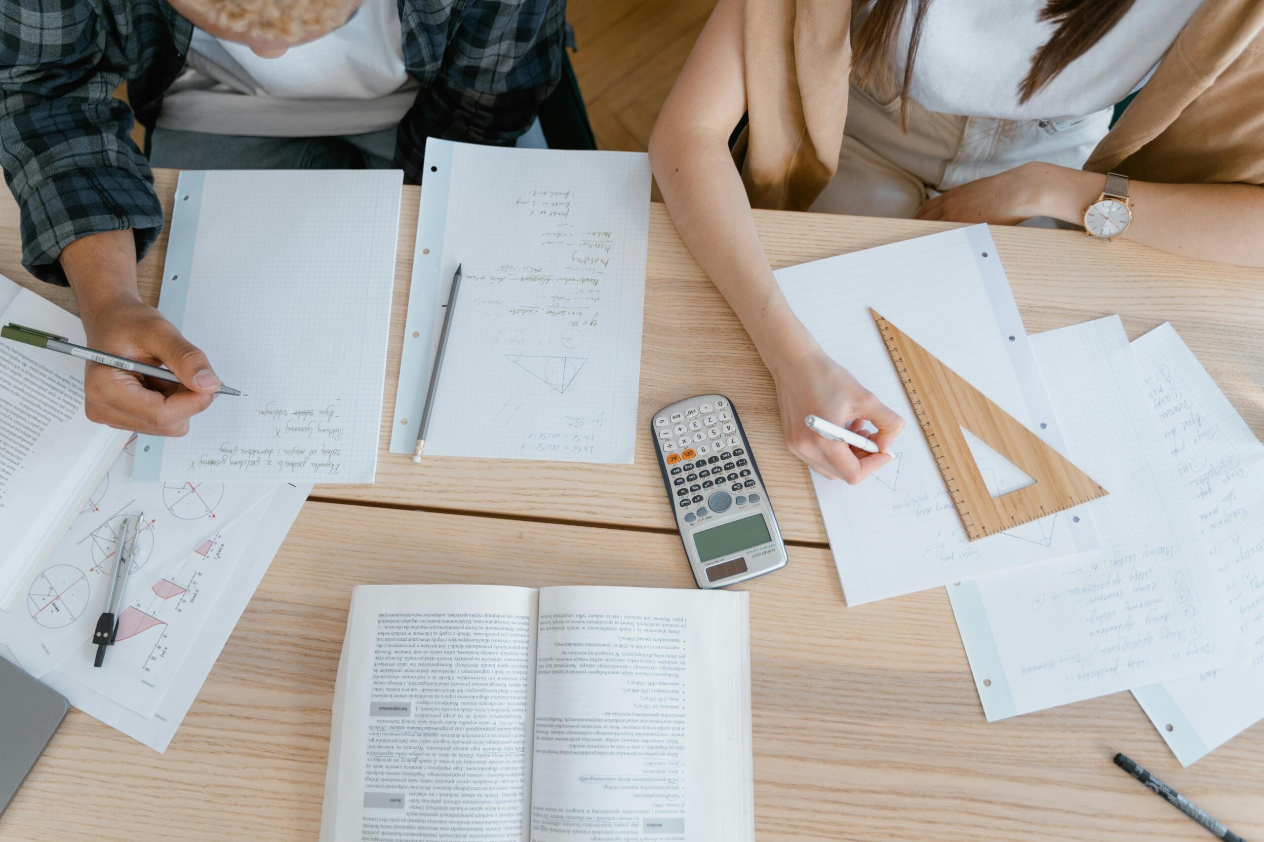 A top‑down view shows notebooks with geometric diagrams, a scientific calculator, pencils and a math textbook on a wooden table while students work together.