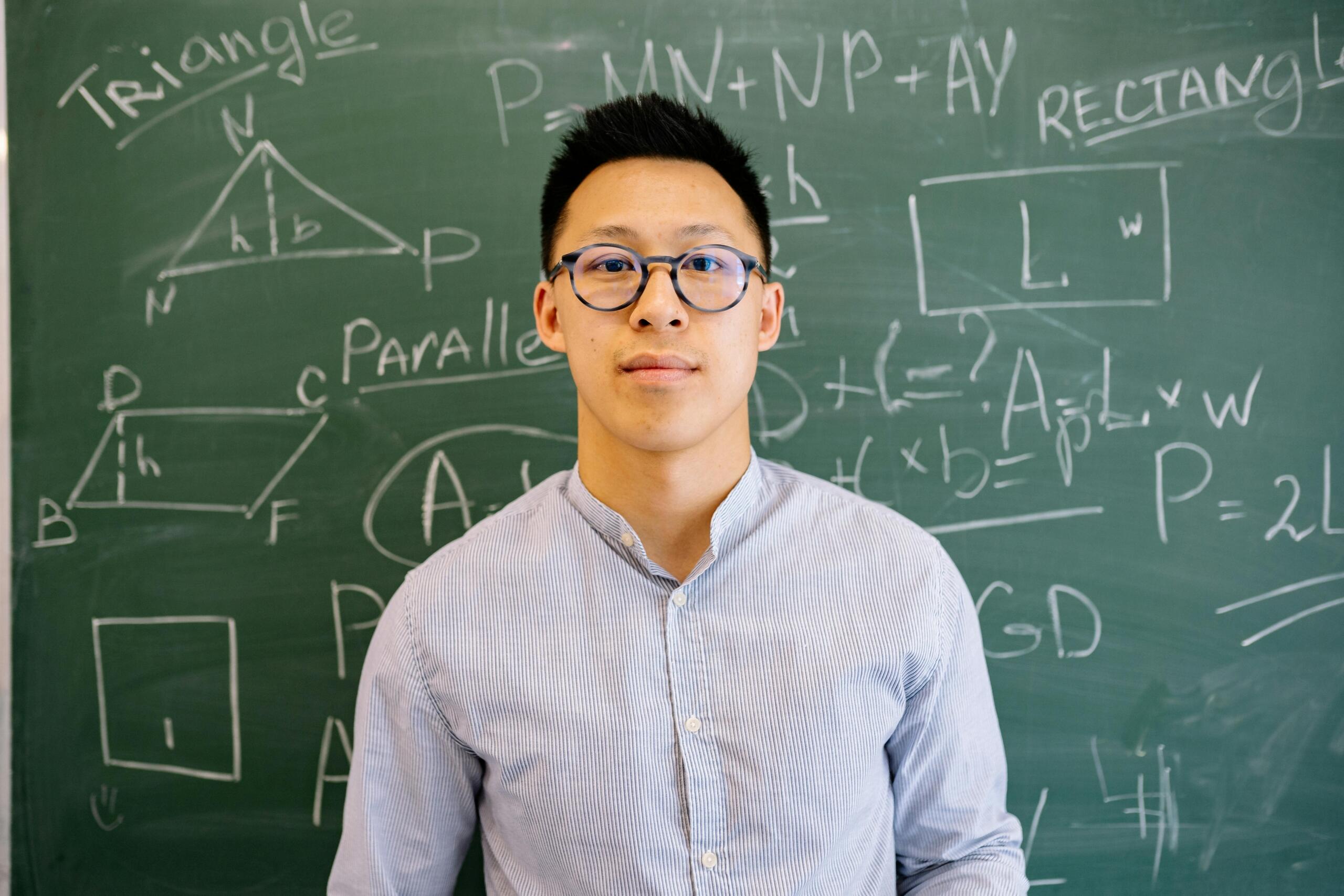 A man wearing glasses and a light blue shirt stands in front of a green chalkboard covered with geometric diagrams and mathematical formulas drawn in white chalk.