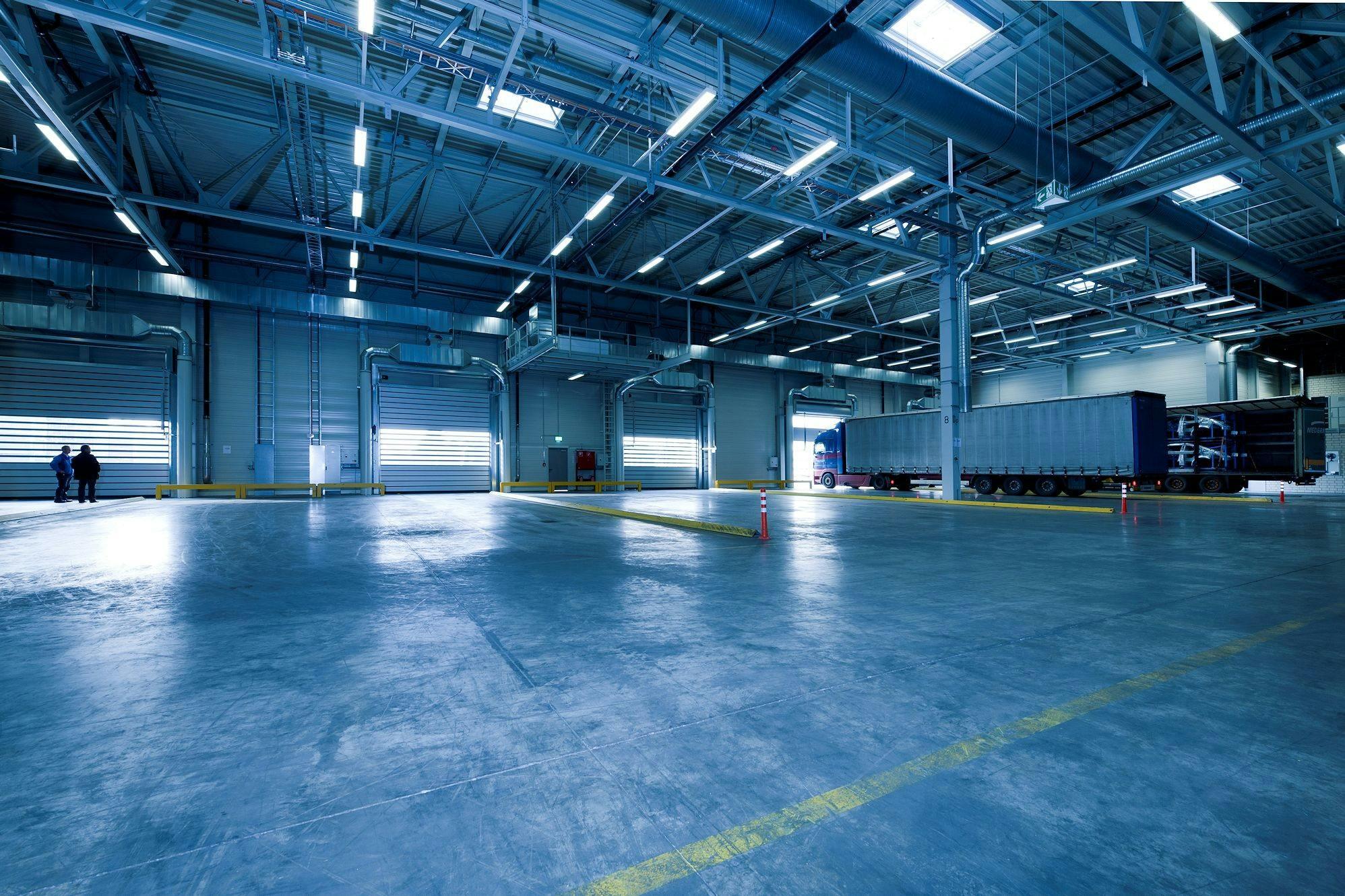The interior of a large empty warehouse shows a polished concrete floor, high metal roof with lights and ducts, loading bay doors and a parked tractor‑trailer; two people stand near one door.
