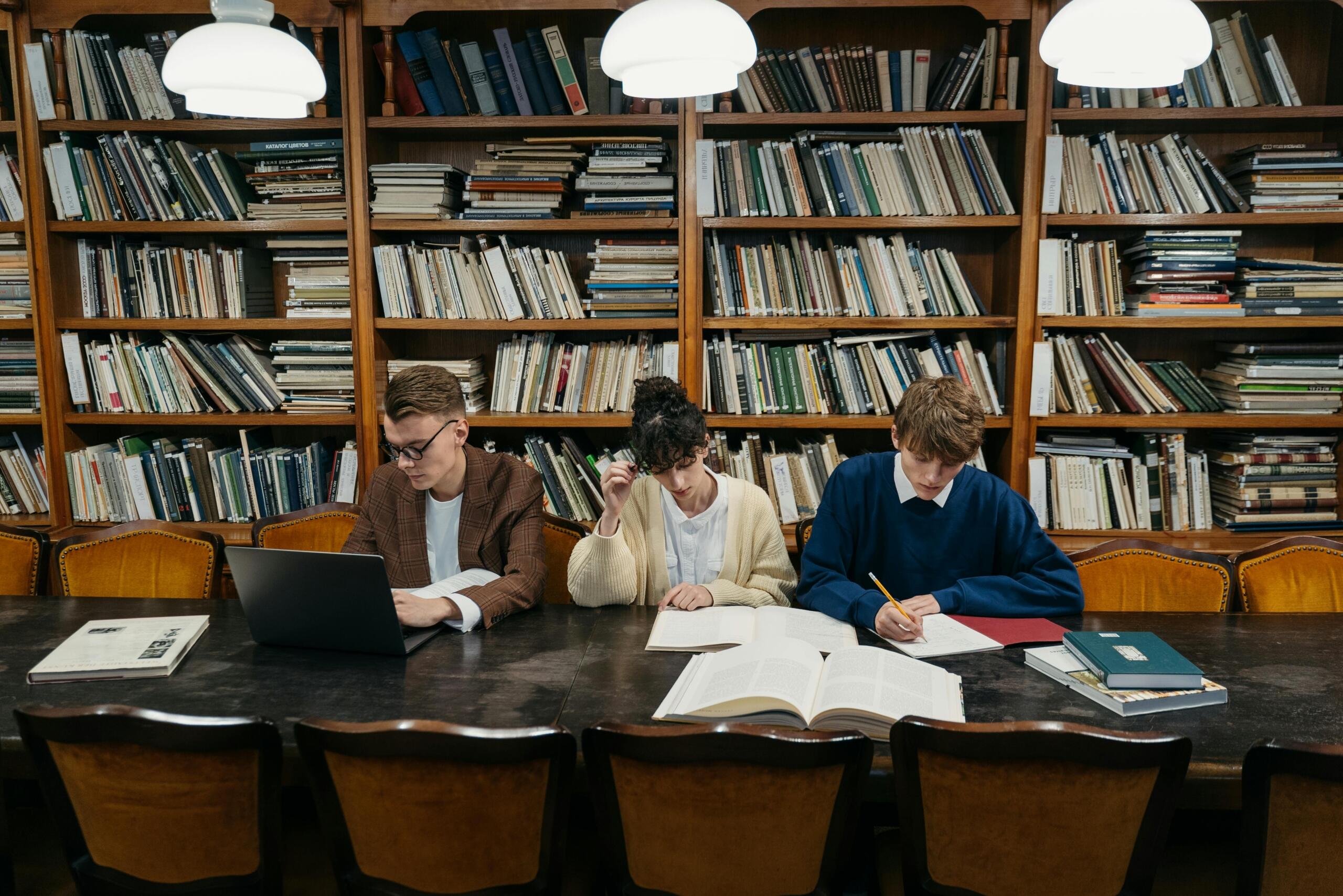 Three university students sit at a large wooden table in a library, surrounded by books and a laptop, with bookshelves in the background.