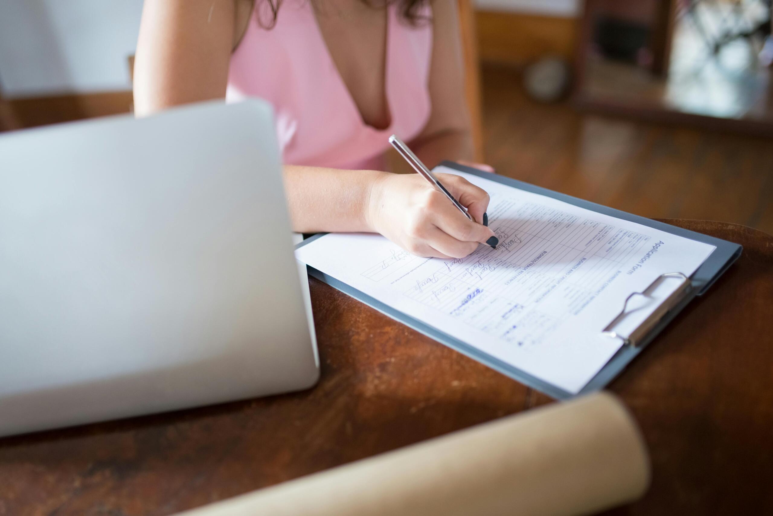 A woman writes on a form attached to a blue clipboard while a laptop, smartphone and papers sit on the desk.