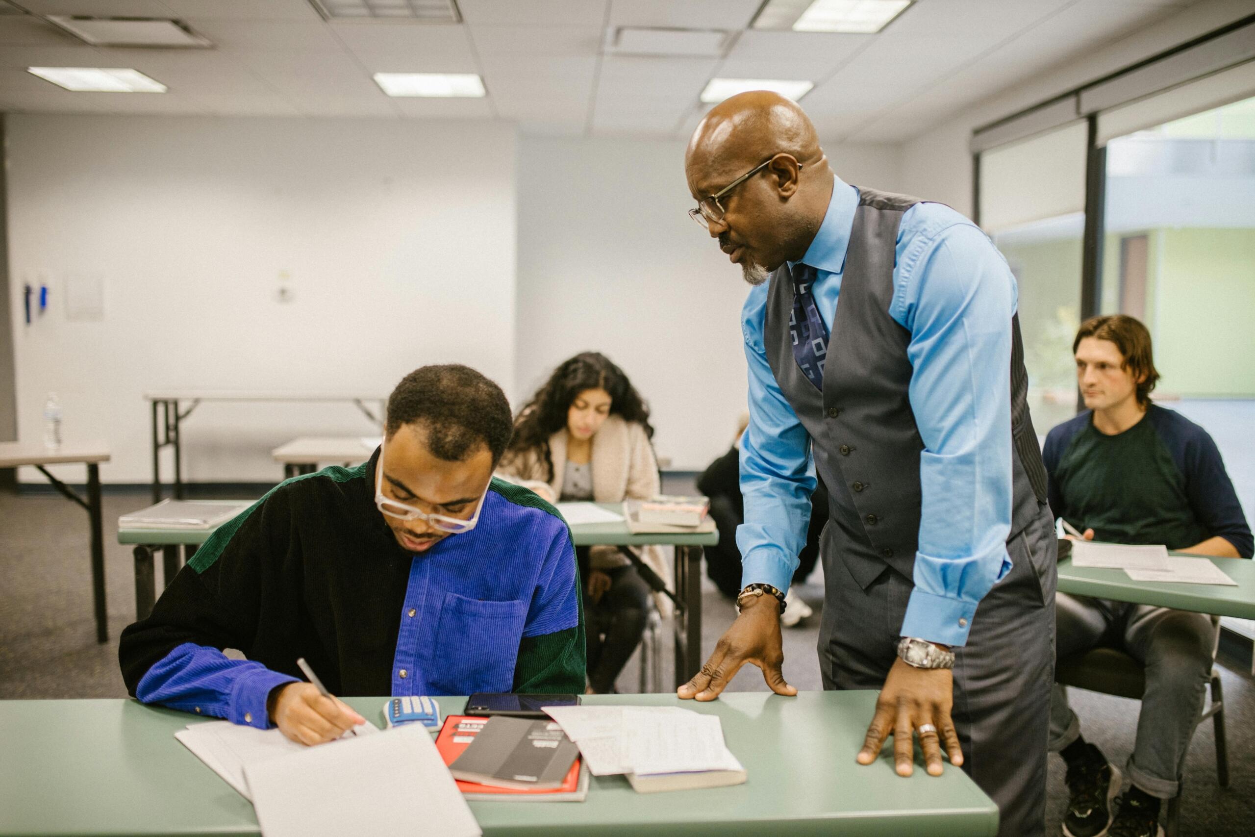 A teacher wearing a waistcoat leans over to assist a student who is writing during an exam in a classroom with other students.