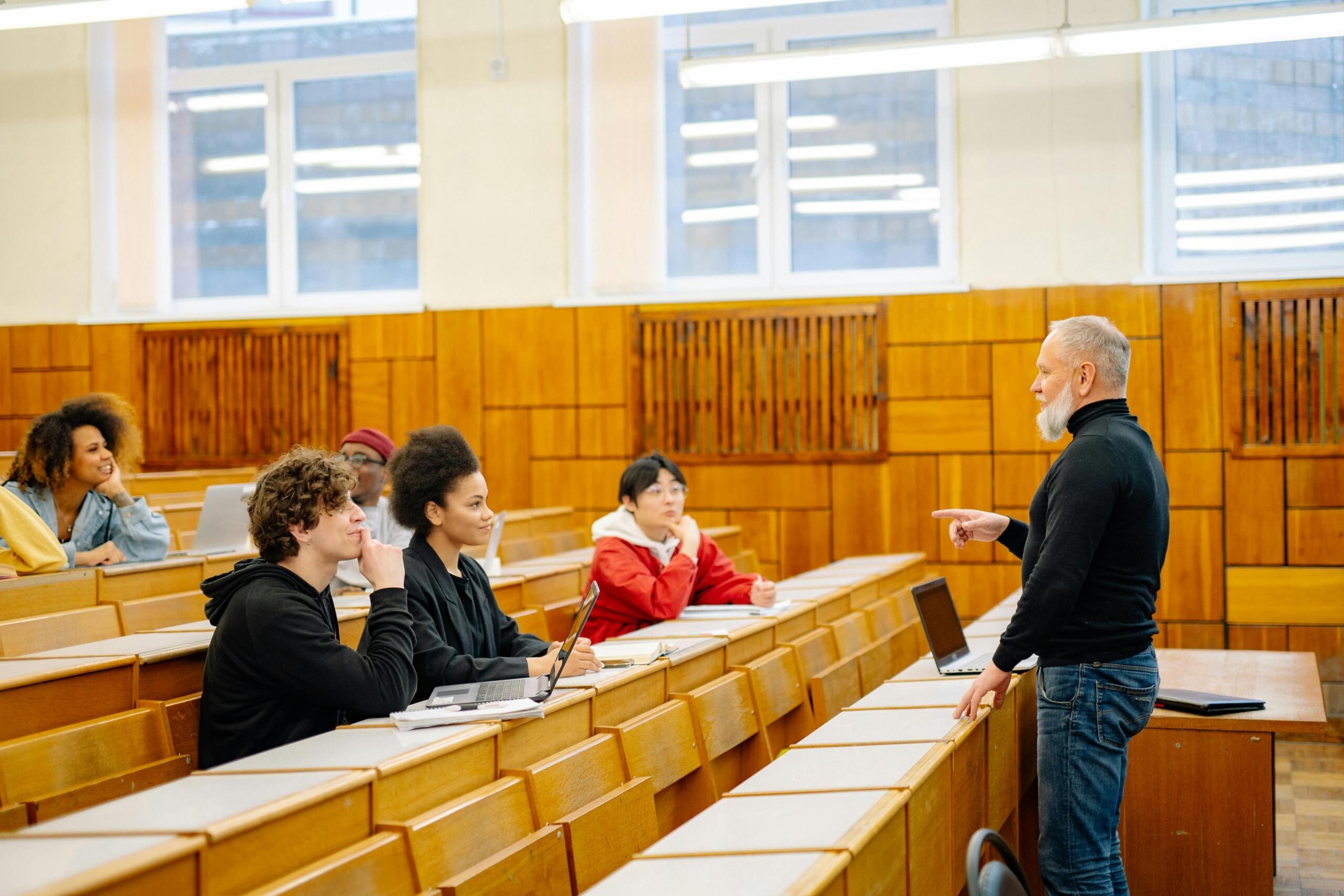 A university lecturer stands at the front of a wood‑panelled lecture hall, addressing rows of seated students taking notes.