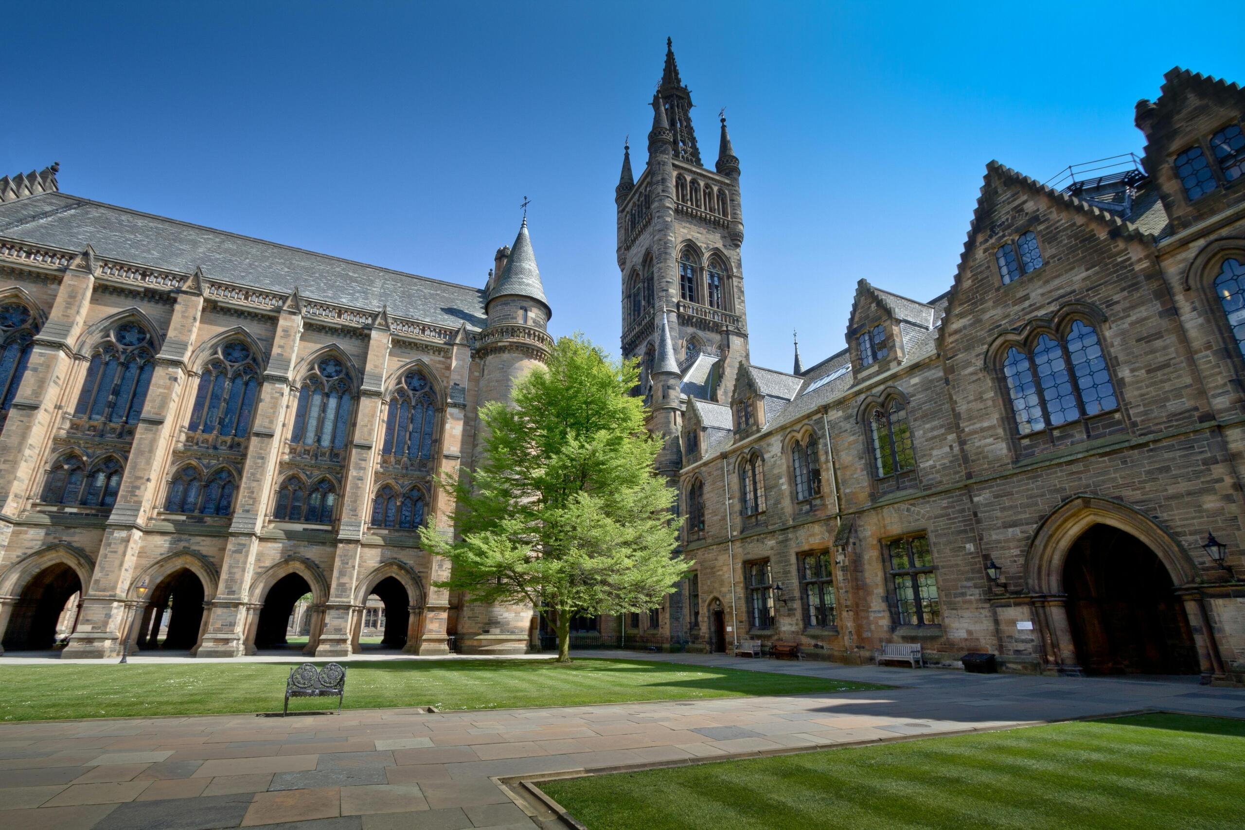 A grand historic university courtyard with a grassy quadrangle is surrounded by arched stone corridors and a central spire under a clear sky.
