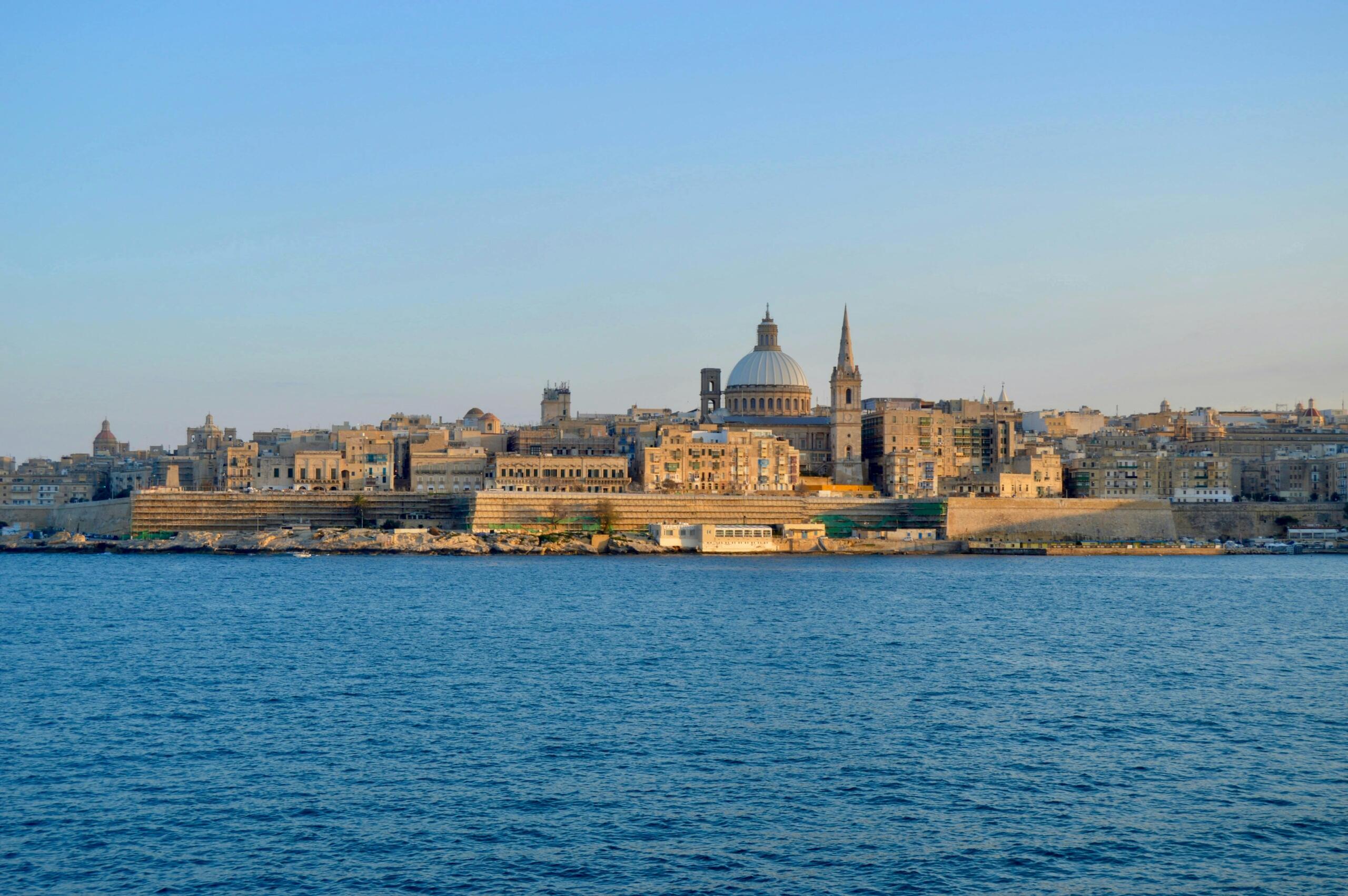 Panoramic view across the harbor towards the historic skyline of Valletta, with a domed cathedral and spire under a blue sky.
