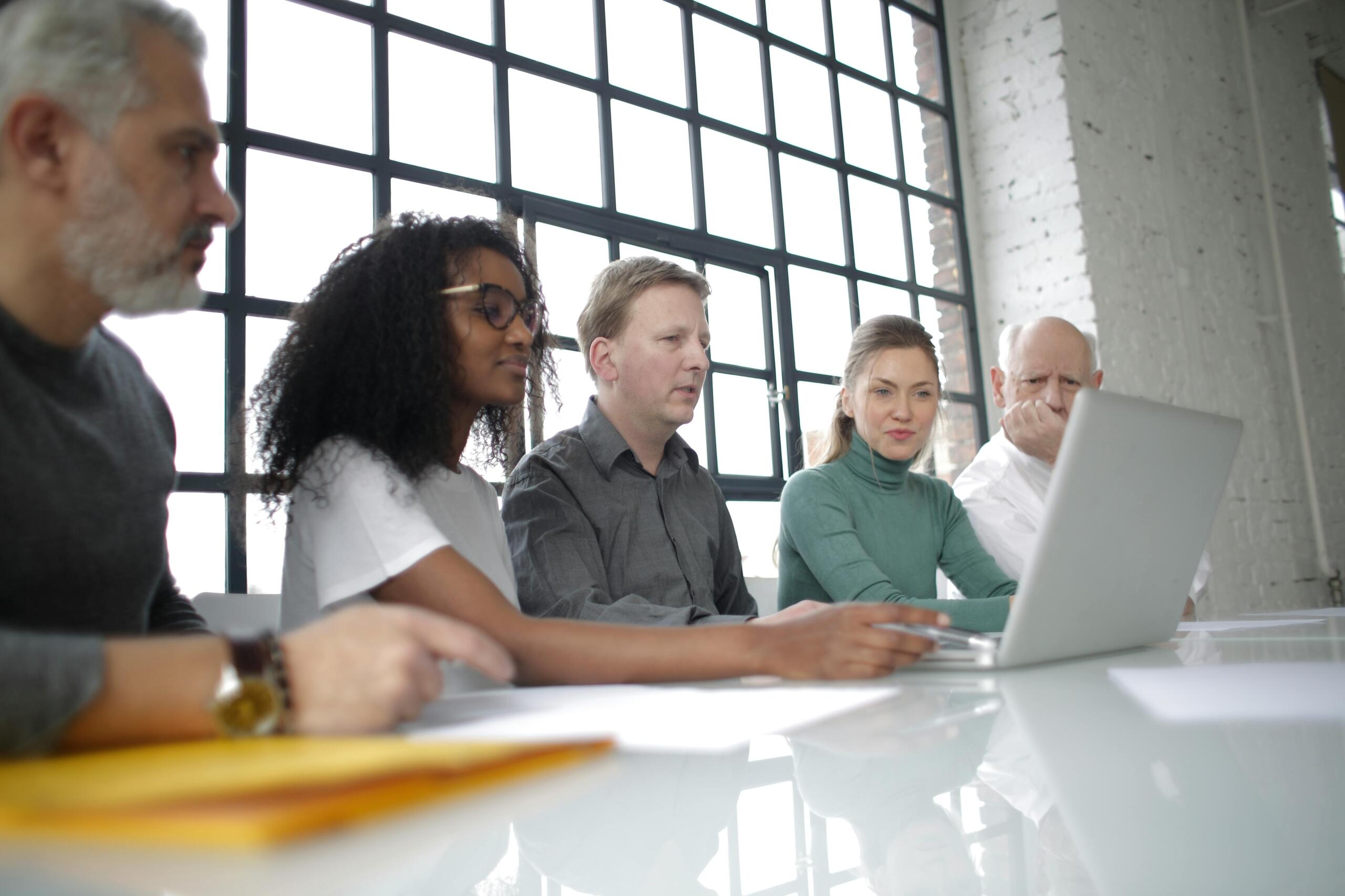 Four professionals of different ages and backgrounds sit at a table in a bright office, focused on a laptop screen.