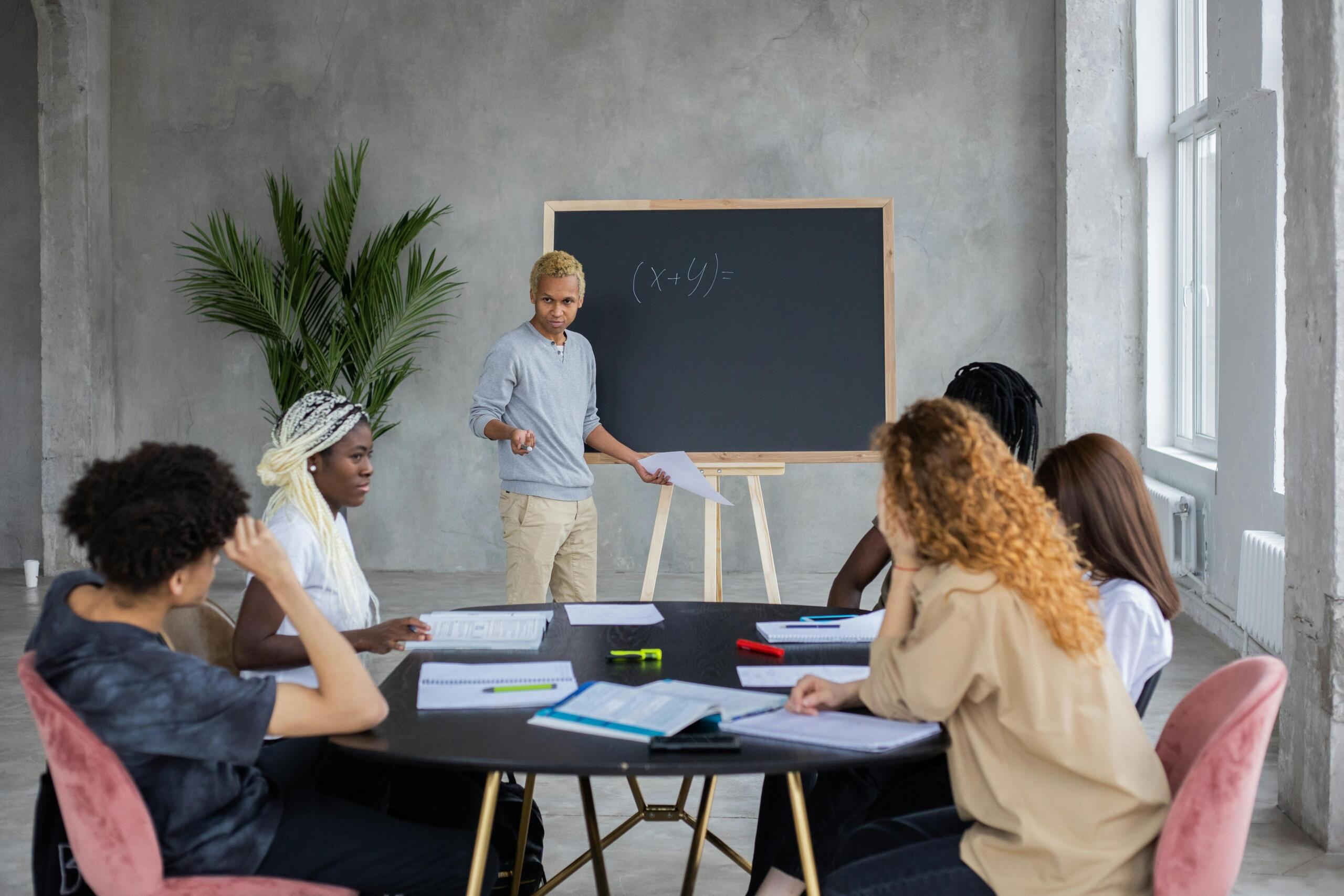 A diverse group of university students sits around a round table with notebooks and laptops while a lecturer writes an equation on a chalkboard.