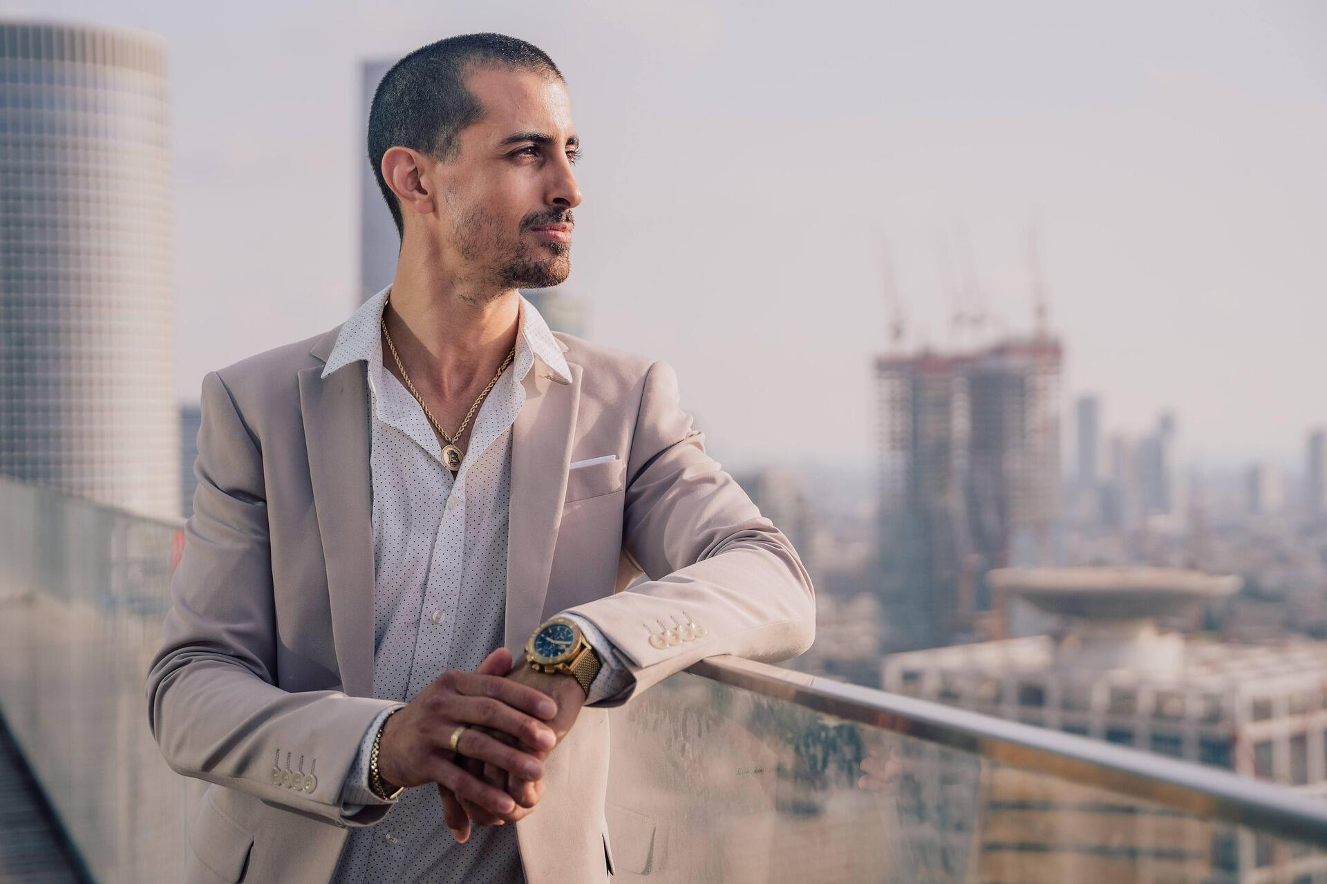 A well-dressed man in a light beige suit standing on a rooftop terrace, looking into the distance with a city skyline and high-rise buildings in the background