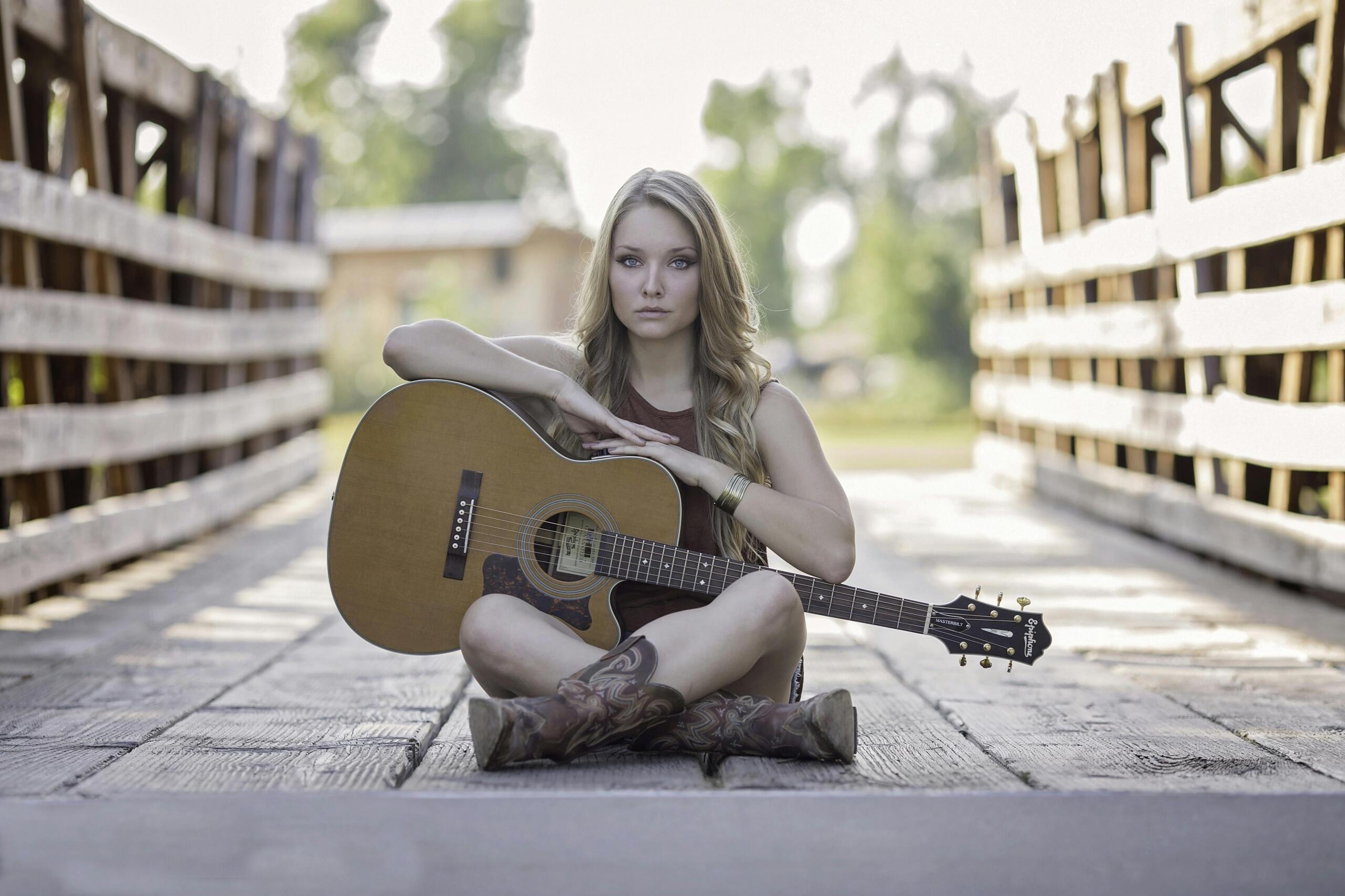 A woman with long blonde hair sits cross‑legged on a wooden bridge outdoors with an acoustic guitar resting on her knees.
