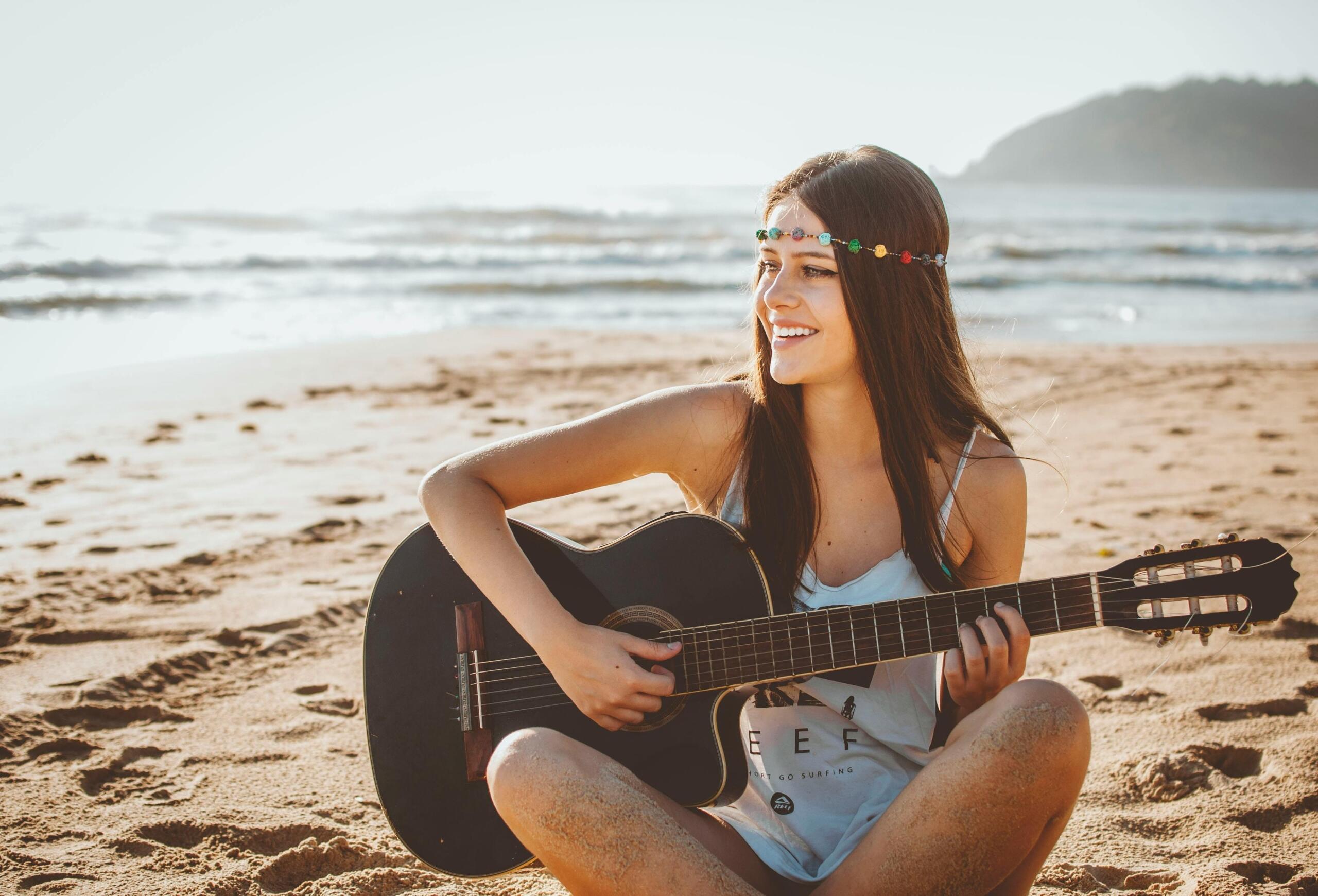 A person is sitting on a sandy beach near the shoreline, holding an acoustic guitar while smiling and looking to the side.