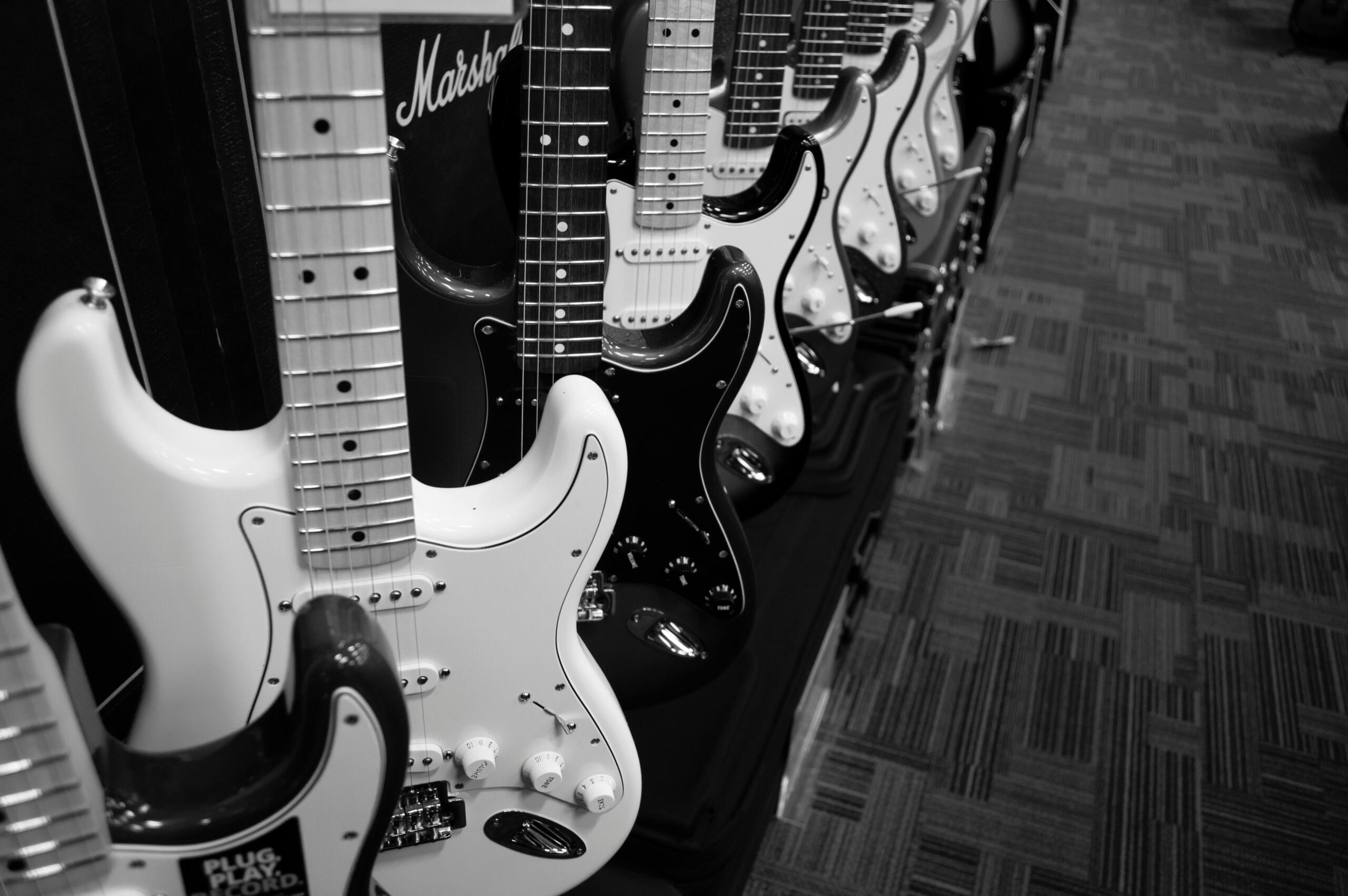 A row of electric guitars is displayed in a shop, with multiple Stratocaster-style models lined up closely together in black and white.