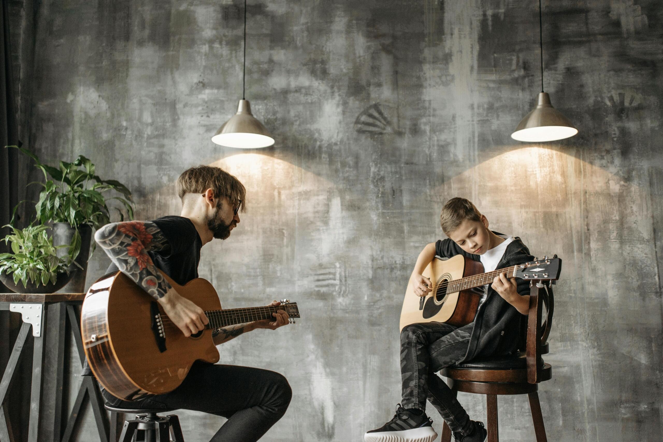 A tattooed male instructor and a boy with an acoustic guitar sit facing each other in a studio with industrial decor and pendant lights.