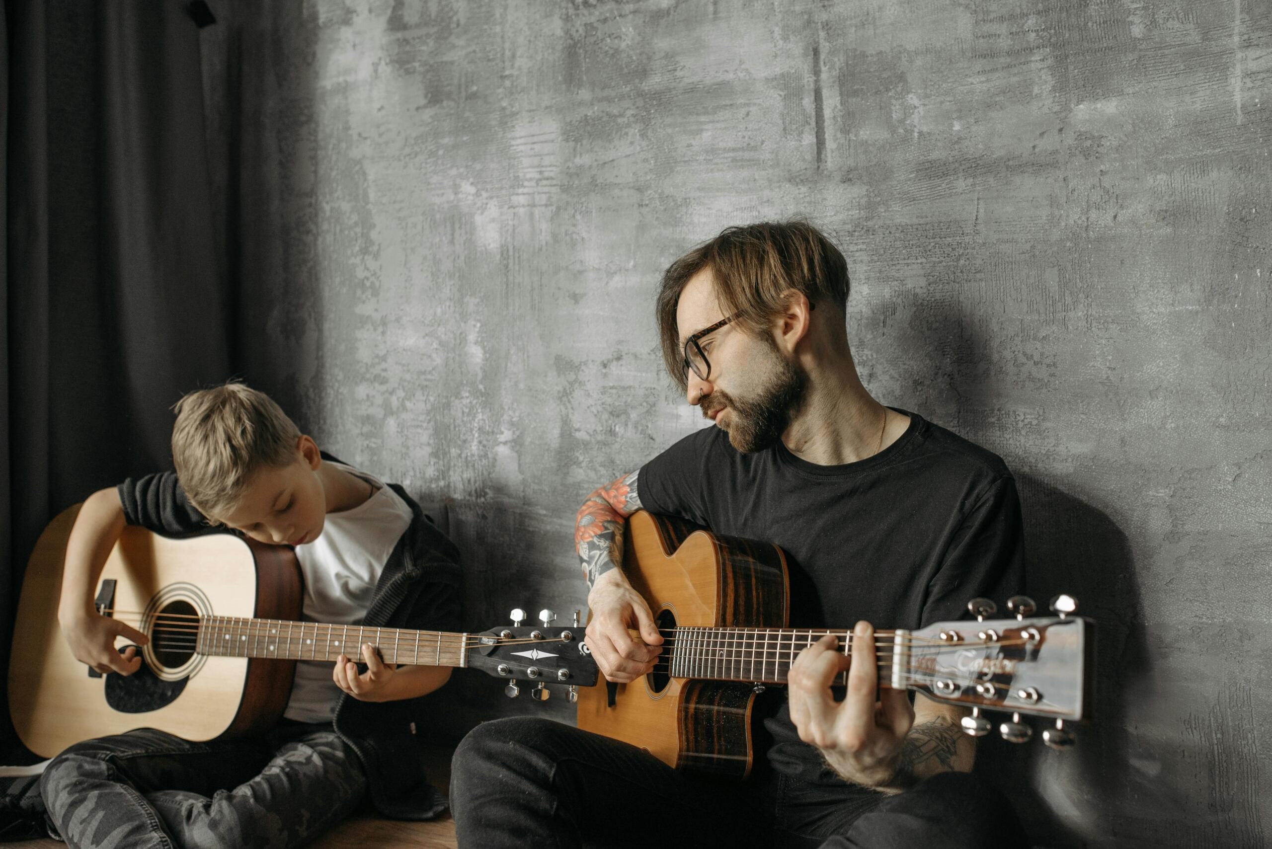 A tattooed adult sits cross‑legged on the floor opposite a young boy; both hold an acoustic guitar and focus on playing together against a gray wall.