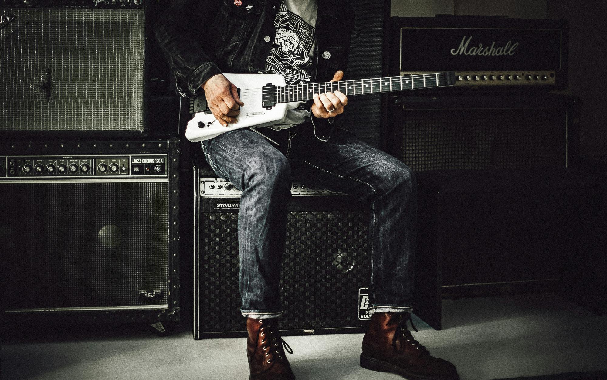 A musician in jeans and boots sits on a guitar amplifier with a white electric guitar resting across his lap; behind him are stacks of amplifier cabinets.