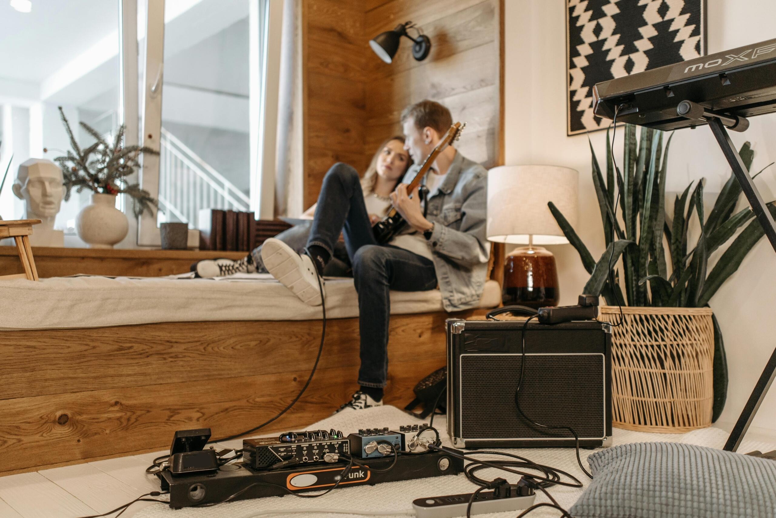 In a modern living room, a young man lies on a raised bed with an electric guitar while another listens; guitar pedals, an amplifier, and a keyboard are in the foreground.