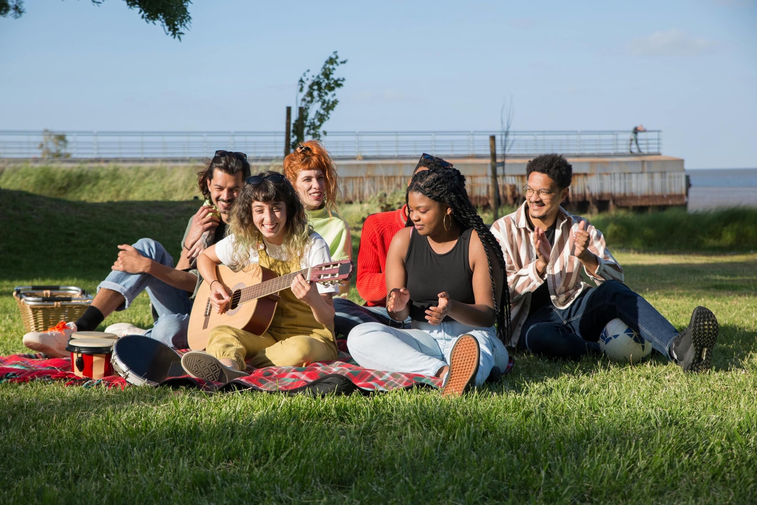 A group of five friends sits on a picnic blanket on a grassy field; one young woman plays an acoustic guitar while the others clap along, with blue sky and water in the background.