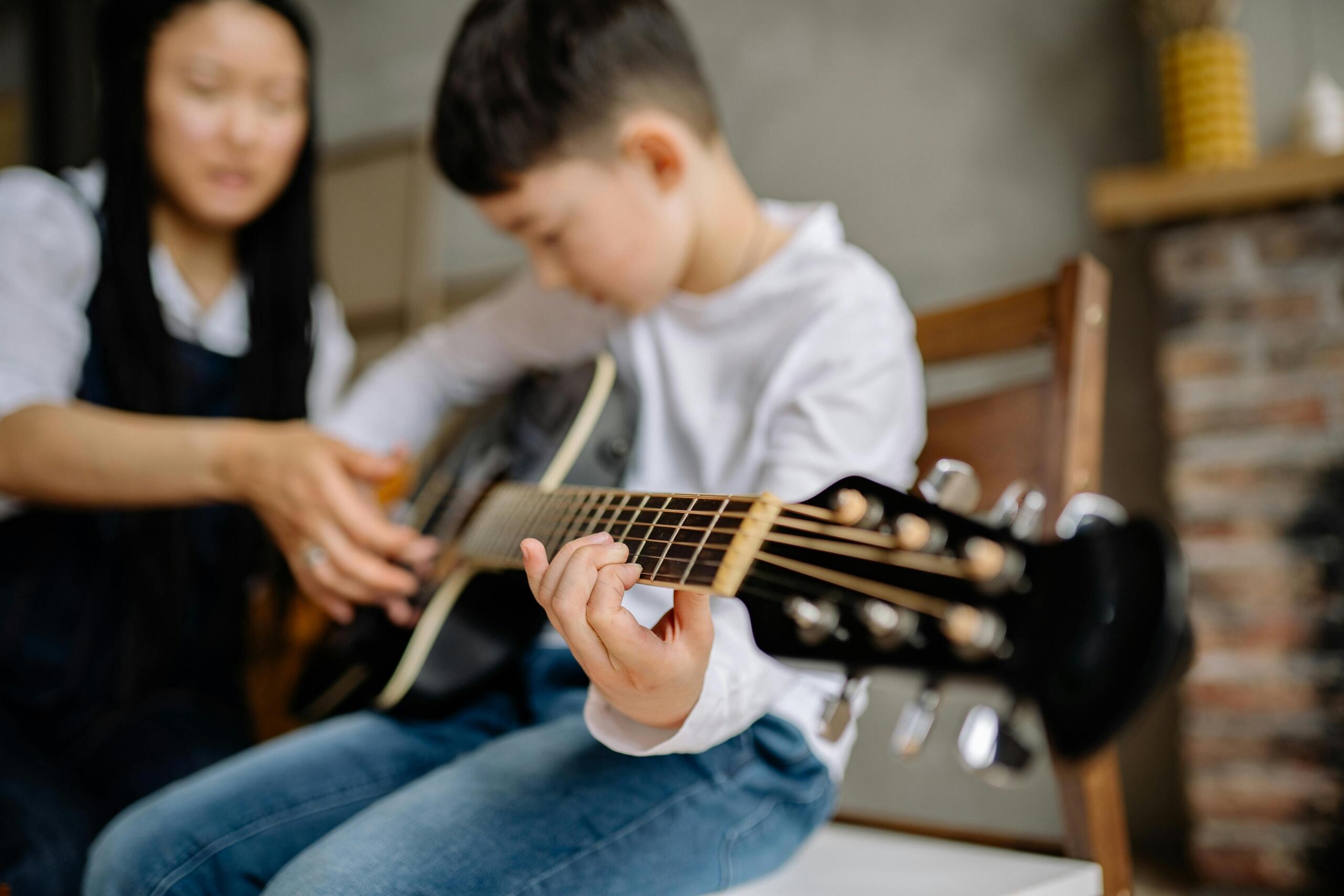 A young boy sits concentrating with an acoustic guitar on his lap; his left hand presses down on the strings while the blurred hand of a tutor next to him provides guidance.