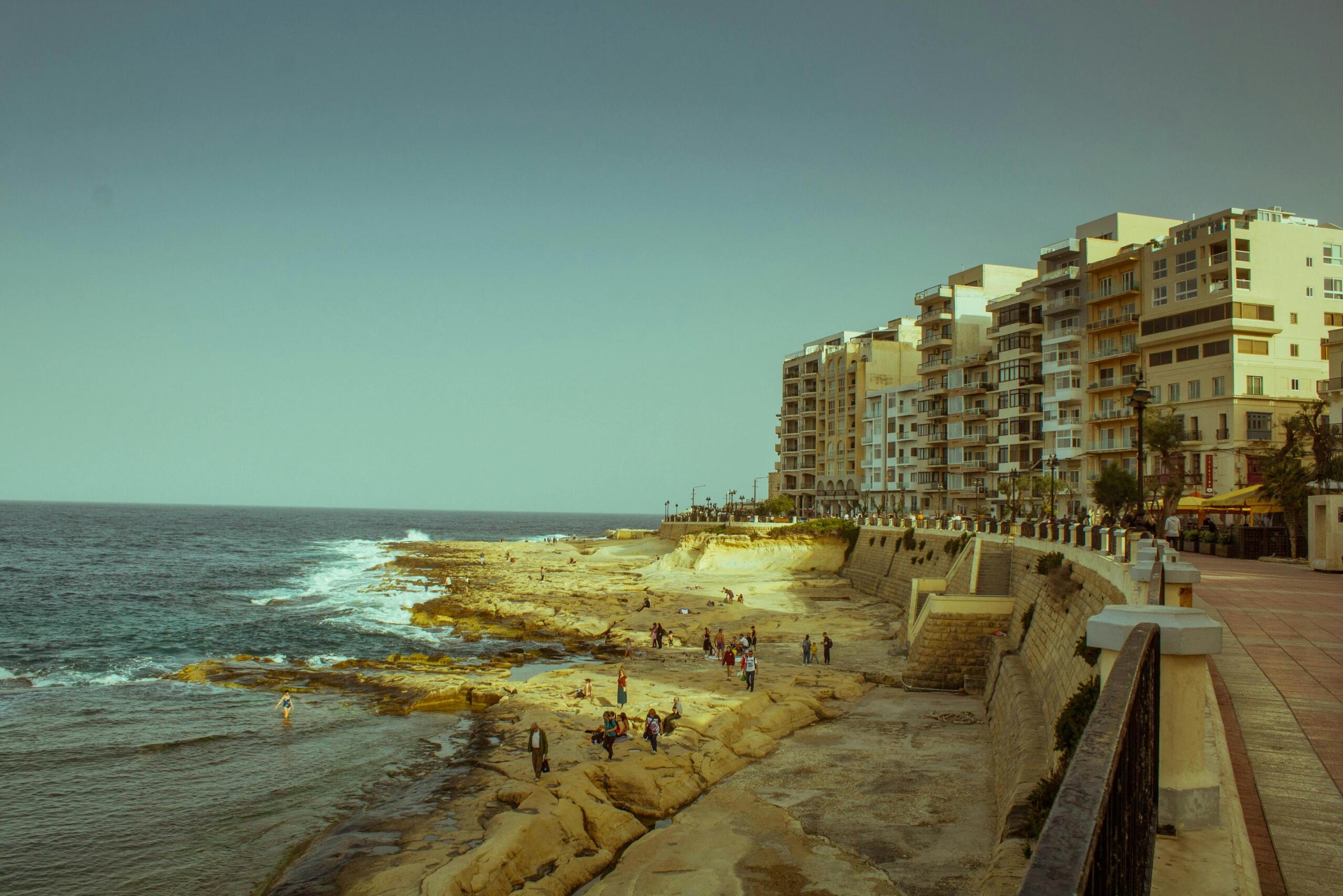 A wide view of Malta’s rocky coastline and promenade with people walking by the sea; modern apartment buildings line the waterfront under a clear sky.