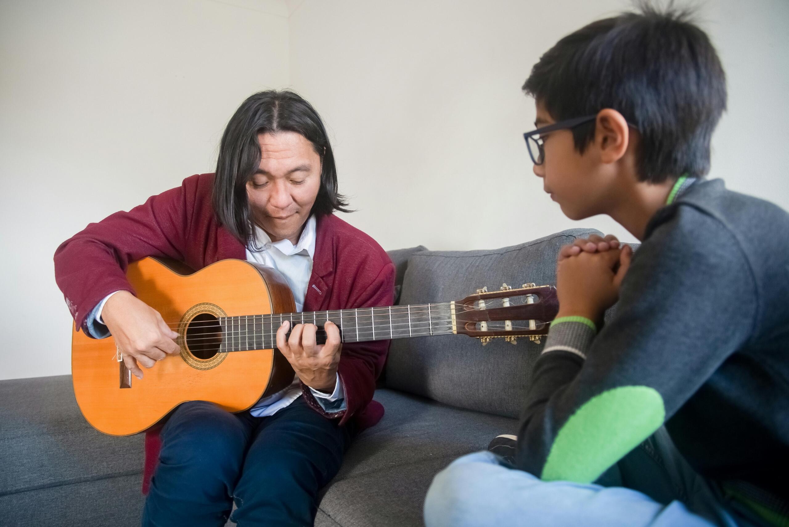 A man with long hair in a red sweater plays a classical guitar while a boy with glasses watches attentively; they sit on a gray sofa in a bright living room.