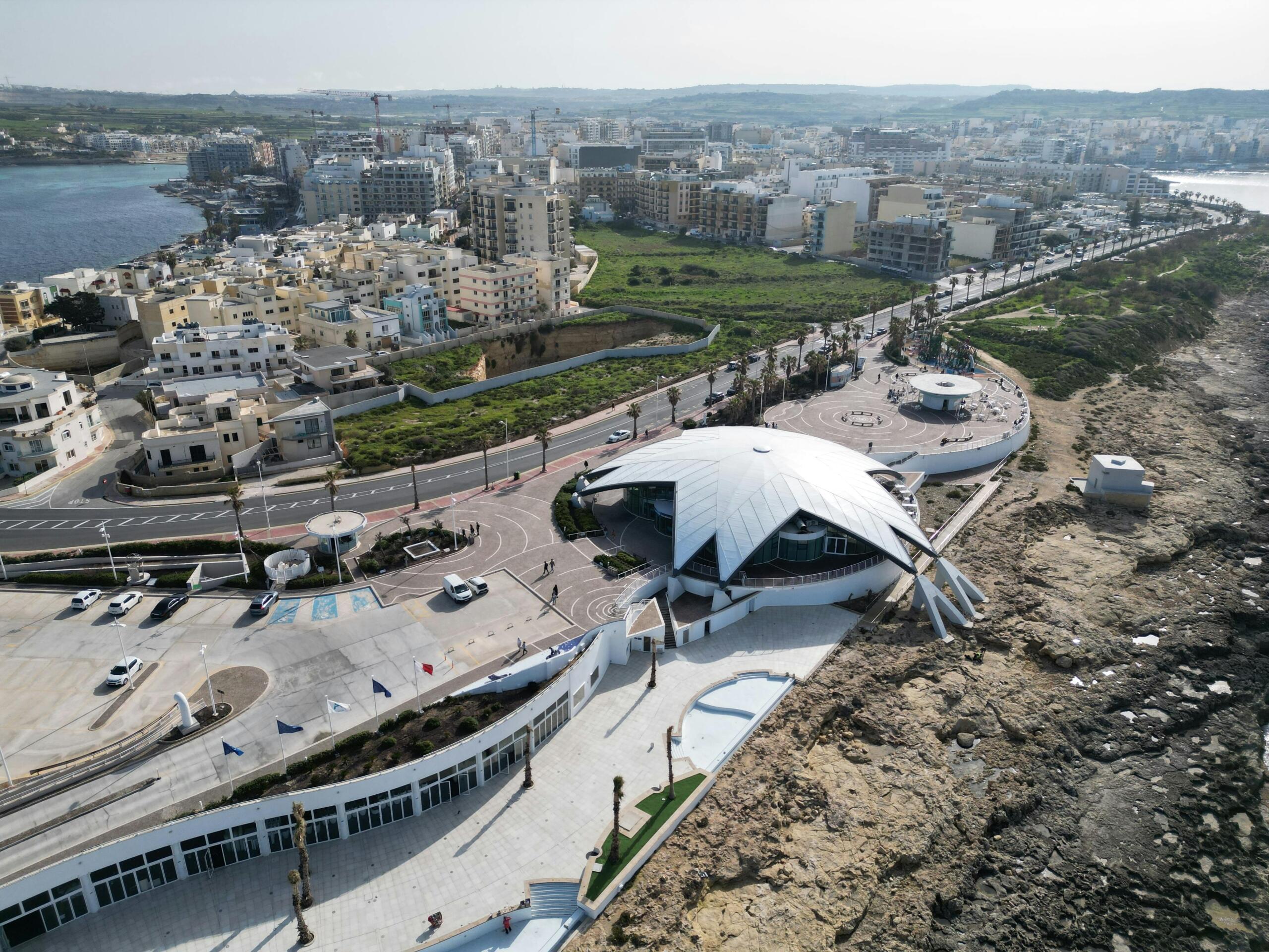 An aerial photo shows St Paul’s Bay with clear blue water, boats drifting, and a mix of white and pastel‑colored buildings along the coast