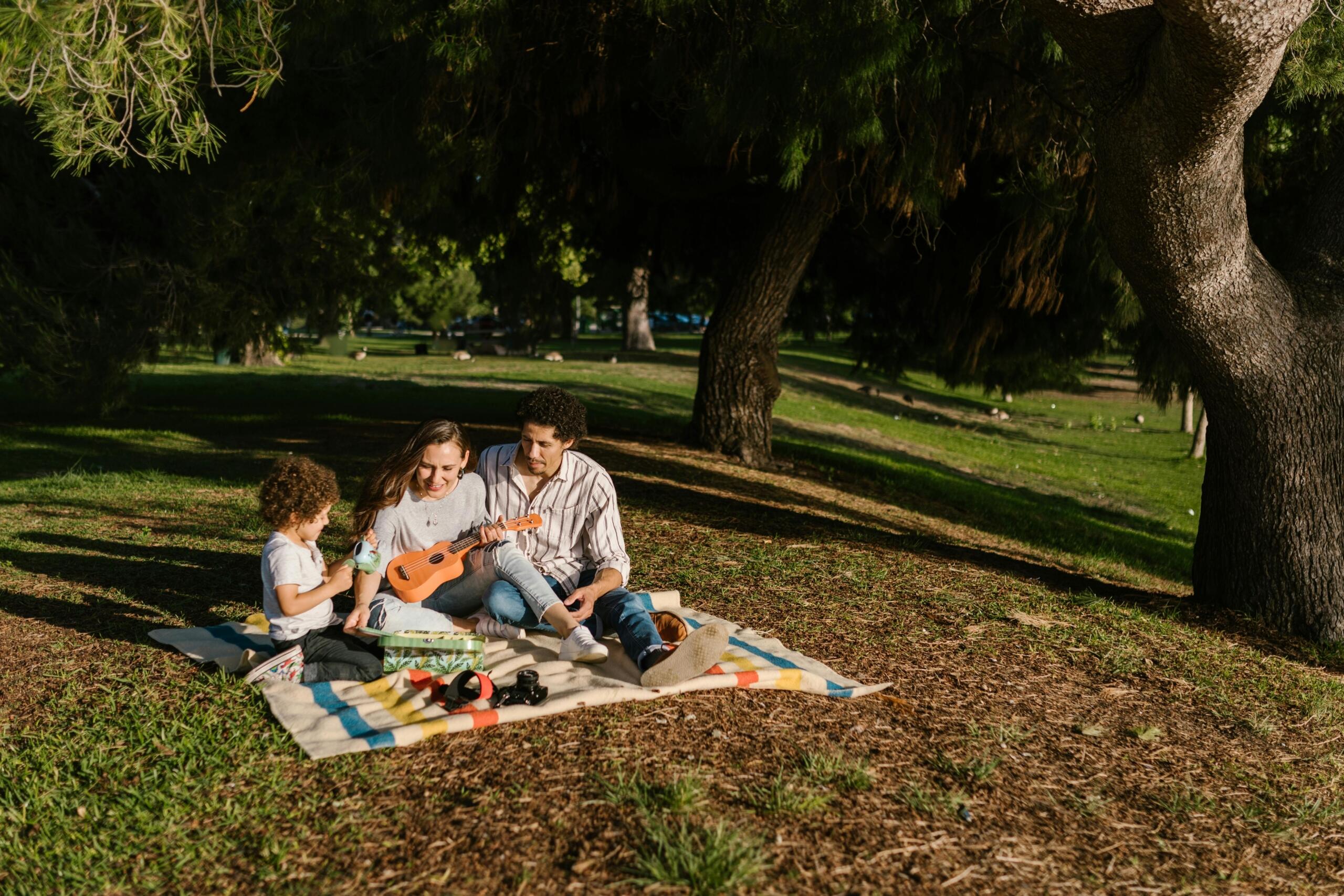 A mother and father sit on a picnic blanket in a park playing a small and a large guitar, while their curly‑haired child watches with a ukulele; sunlight filters through the trees.