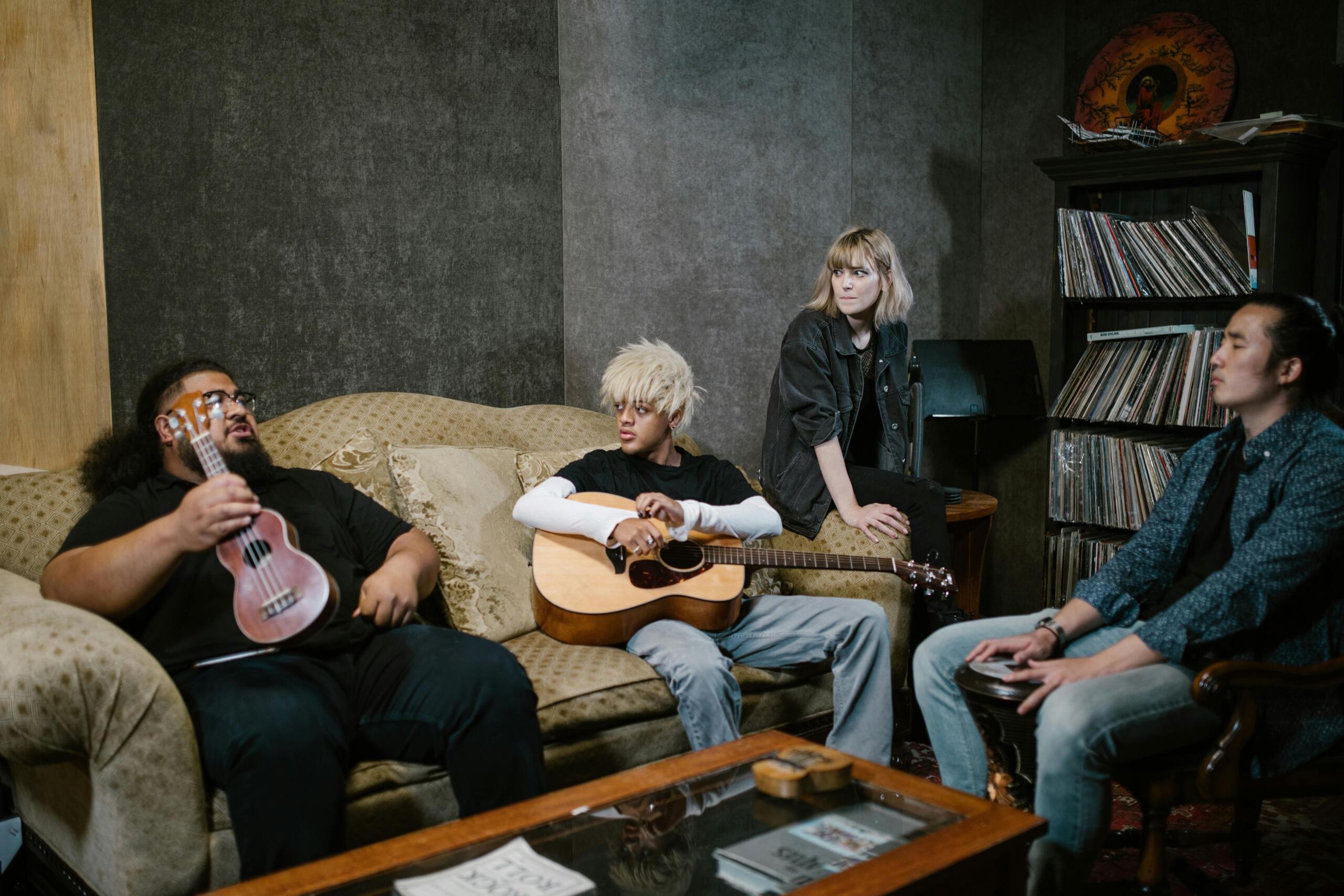 Four young adults sit in a cozy studio: a man with an afro plays a pink ukulele, a blond guitarist strums an acoustic guitar, a woman watches from the armrest, and a man holds a djembe; vintage records and furniture are in the background.