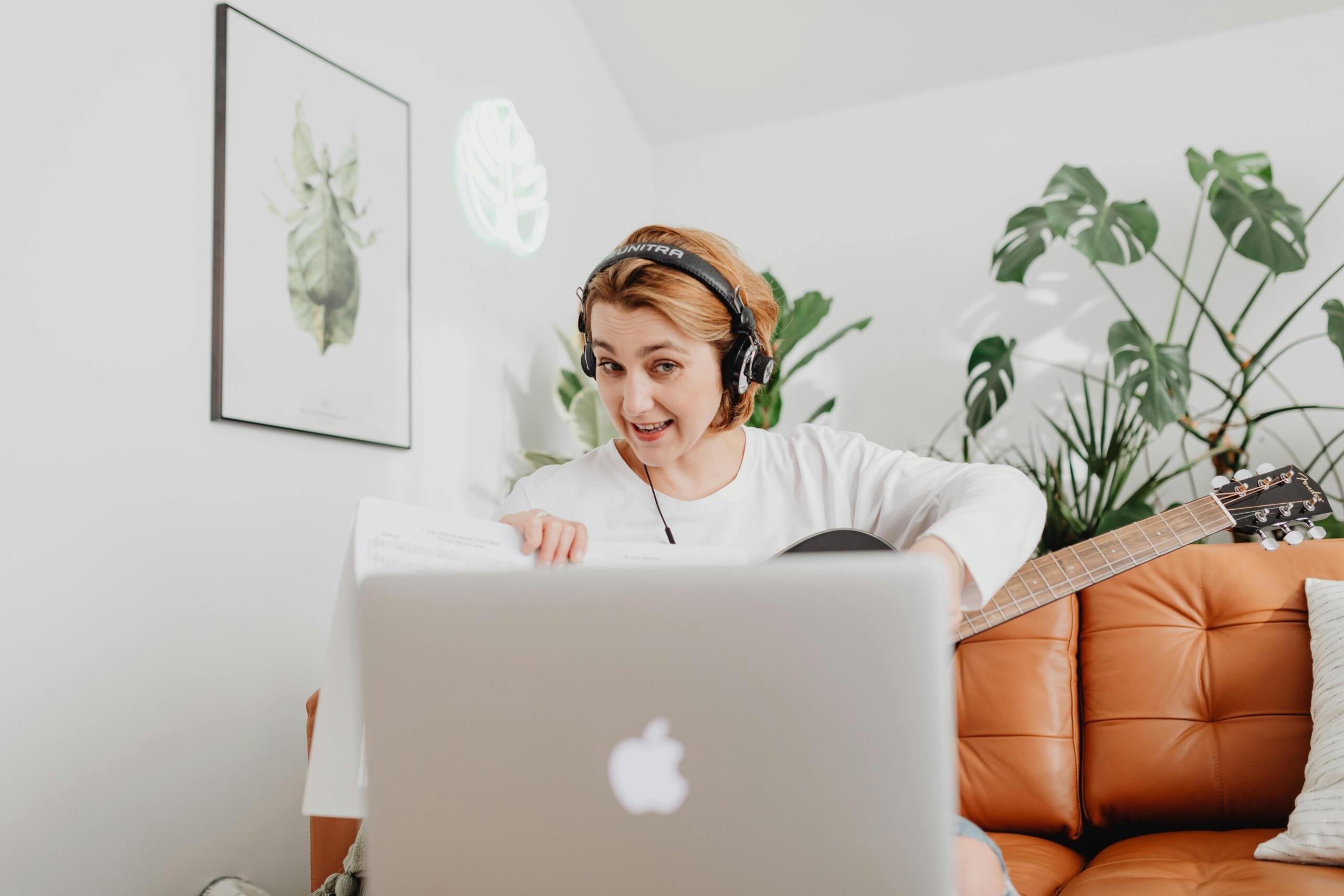 In a modern living room, a young woman sits on a couch with an acoustic guitar on her lap; an open laptop and sheet music are in front of her, and plants fill the room.