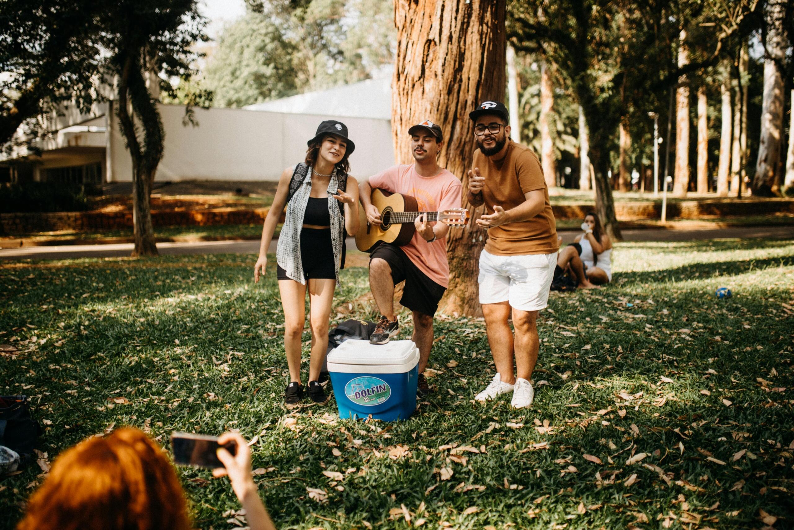 A small group of friends in a park gathers around a guitarist; one man strums while two others sing and everyone smiles, with a cooler in the foreground.