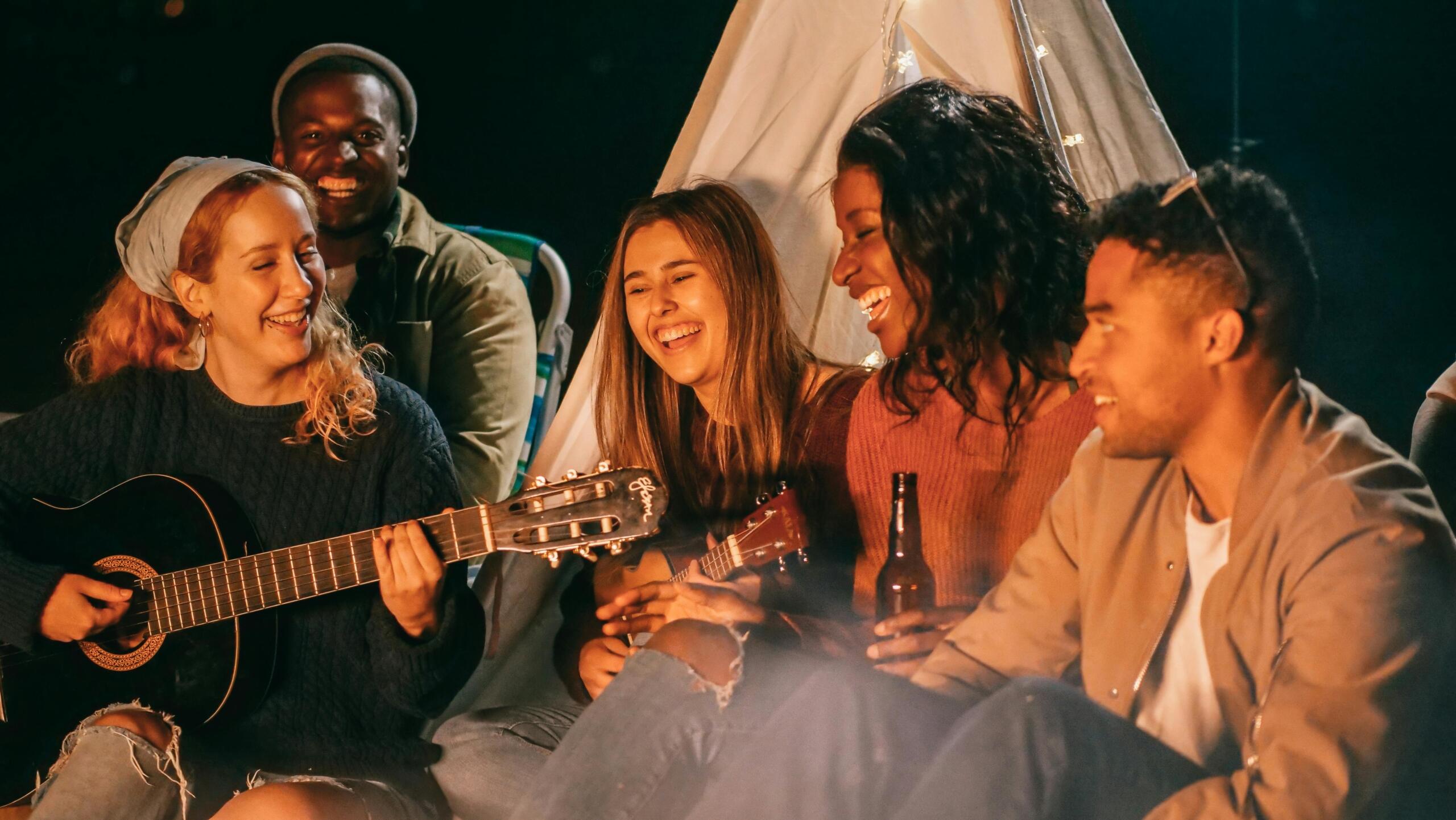 At night on the beach, friends sit on the sand around a tent and campfire; one woman plays a guitar while the others clap, laugh and hold drinks.