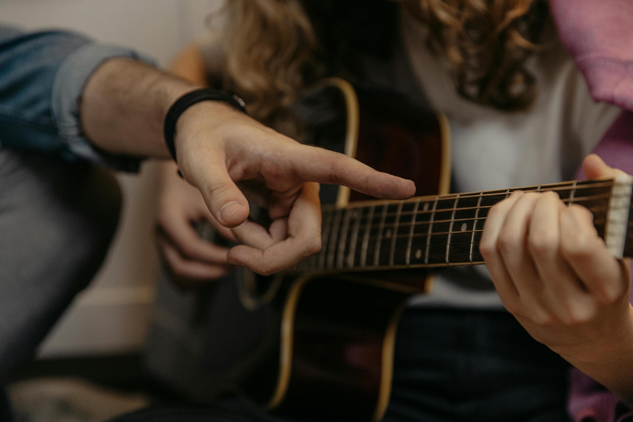 A tutor’s finger points to a specific fret on a student’s acoustic guitar as the student presses down the strings with their left hand.