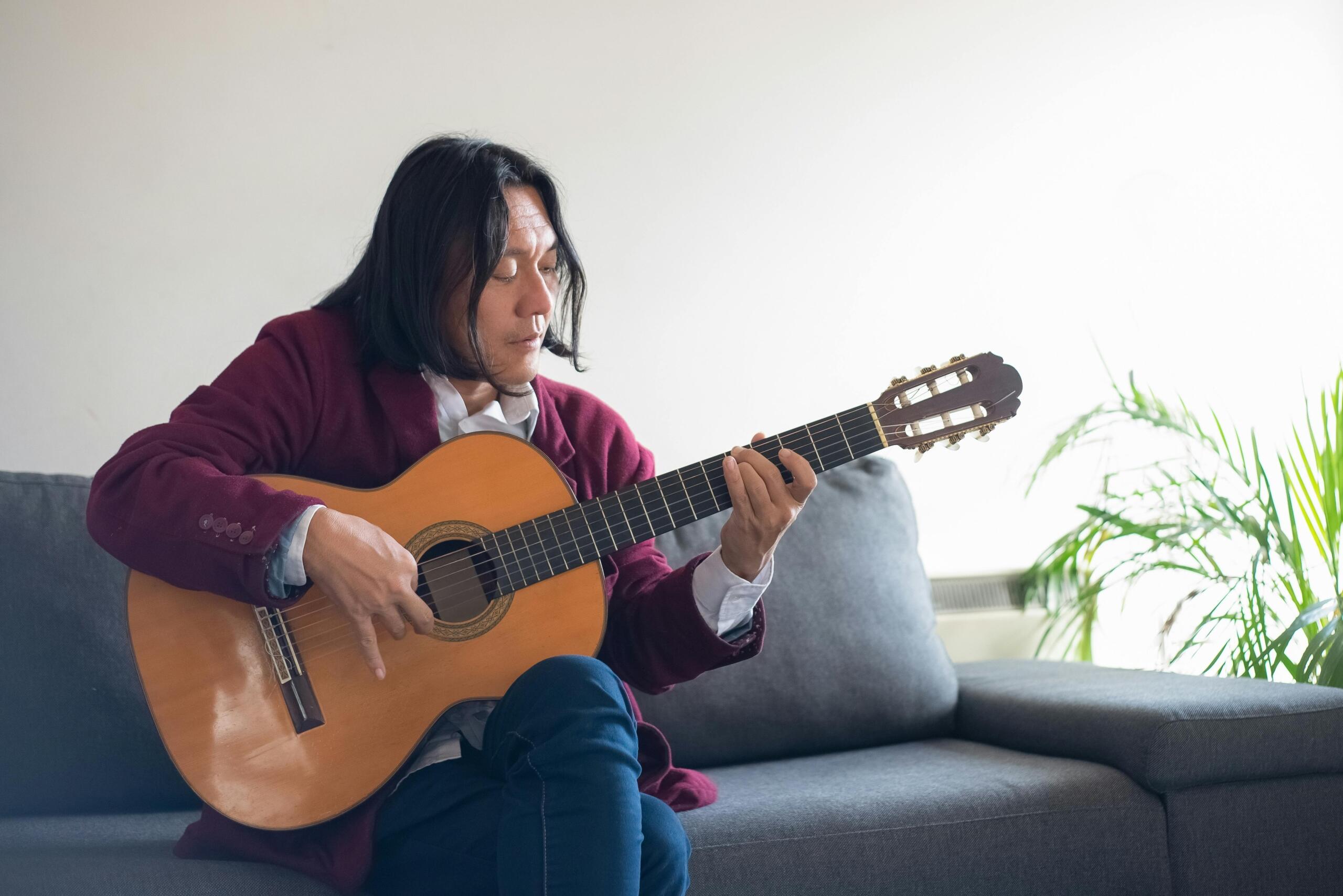A person sitting indoors on a couch, focused on playing an acoustic guitar with both hands positioned on the instrument.