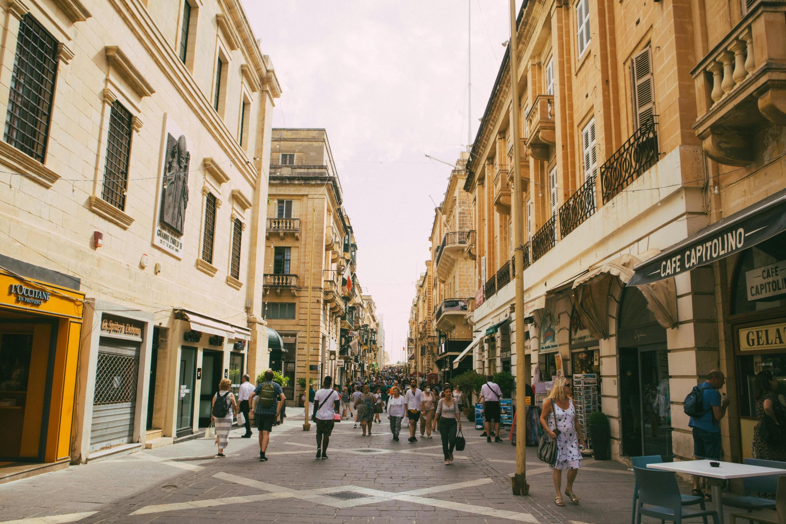 People walking and shopping along Merchant Street in Valletta, Malta, surrounded by historic limestone buildings and lively cafés.