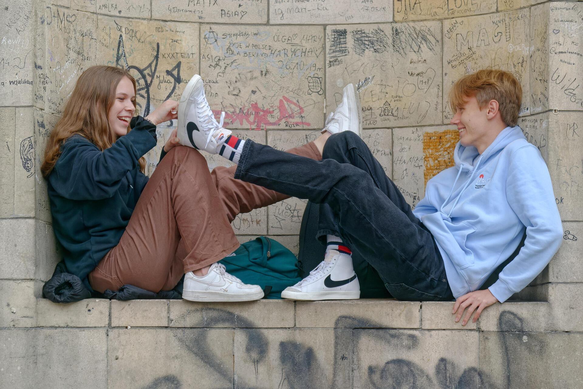 Two teenagers sitting on a stone ledge covered in graffiti, laughing and playfully touching their shoes together while wearing casual hoodies and sneakers.