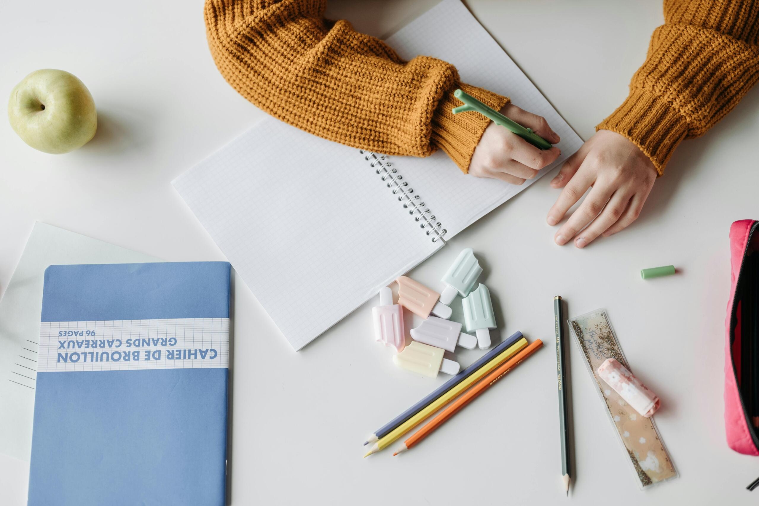 Student writing in a notebook surrounded by pencils, erasers, and a school workbook on a desk.