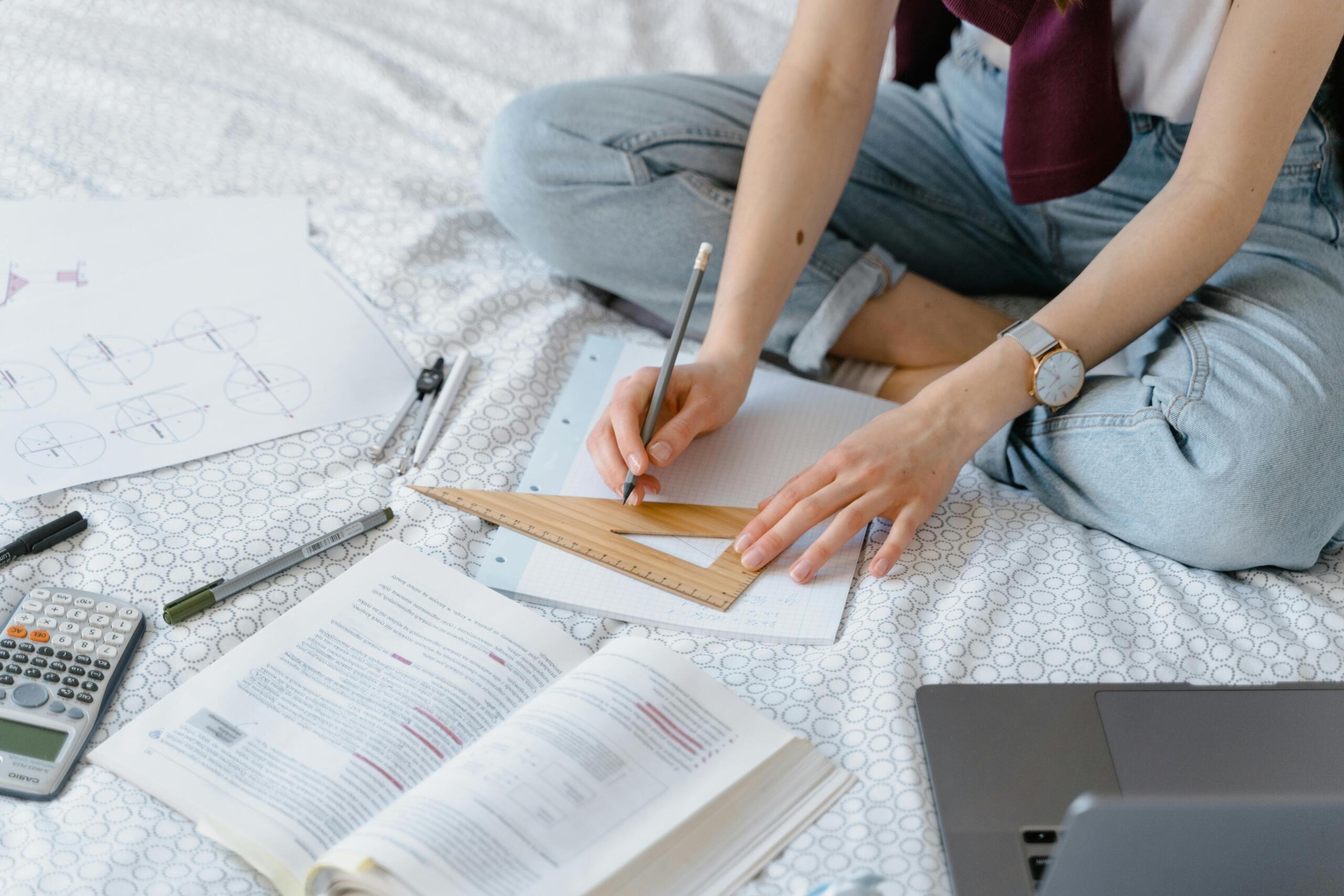 Person sitting on a bed using a ruler and pencil for geometry work with textbooks and tools around them.