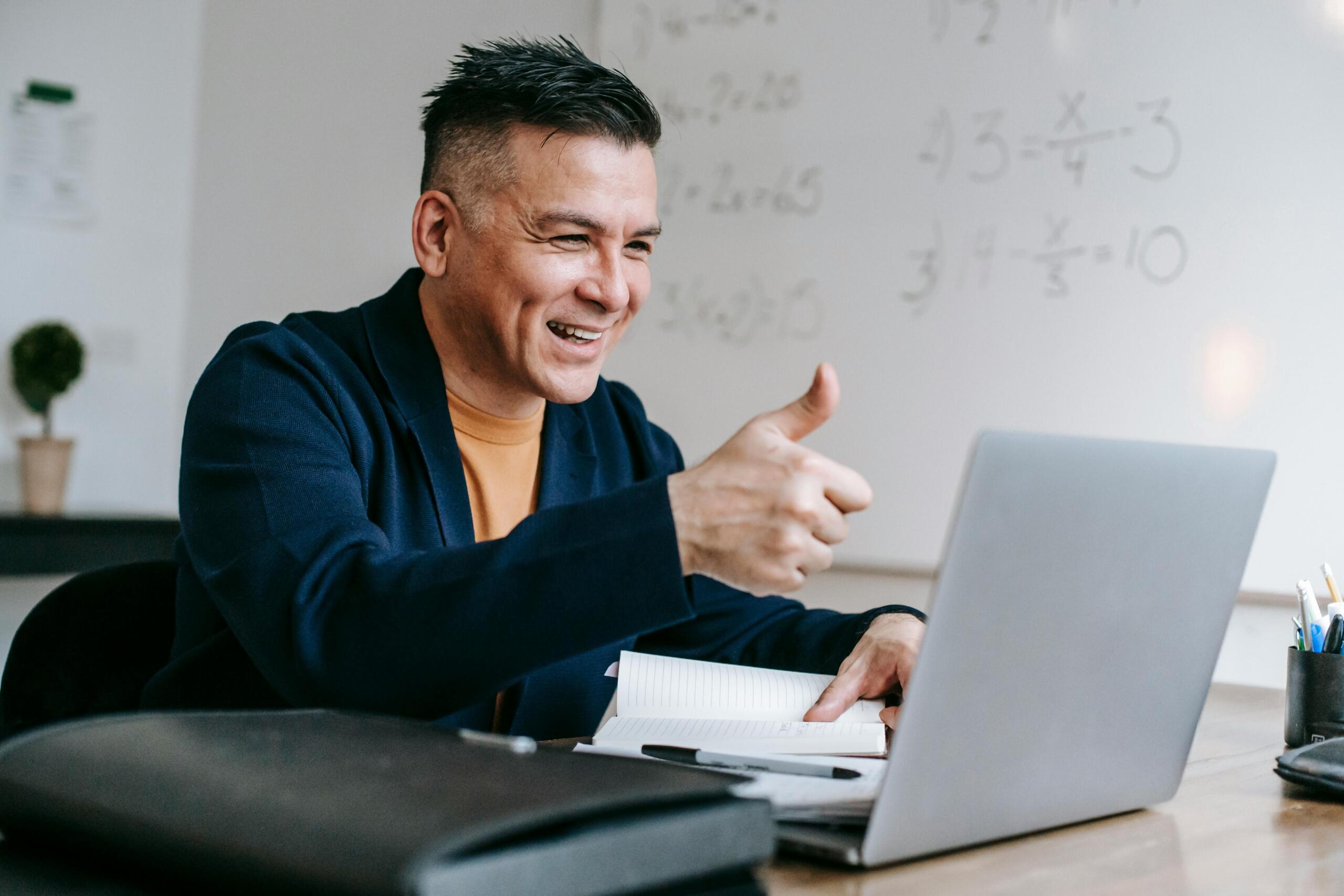Teacher smiling and giving a thumbs up while teaching from a laptop with a whiteboard behind him.