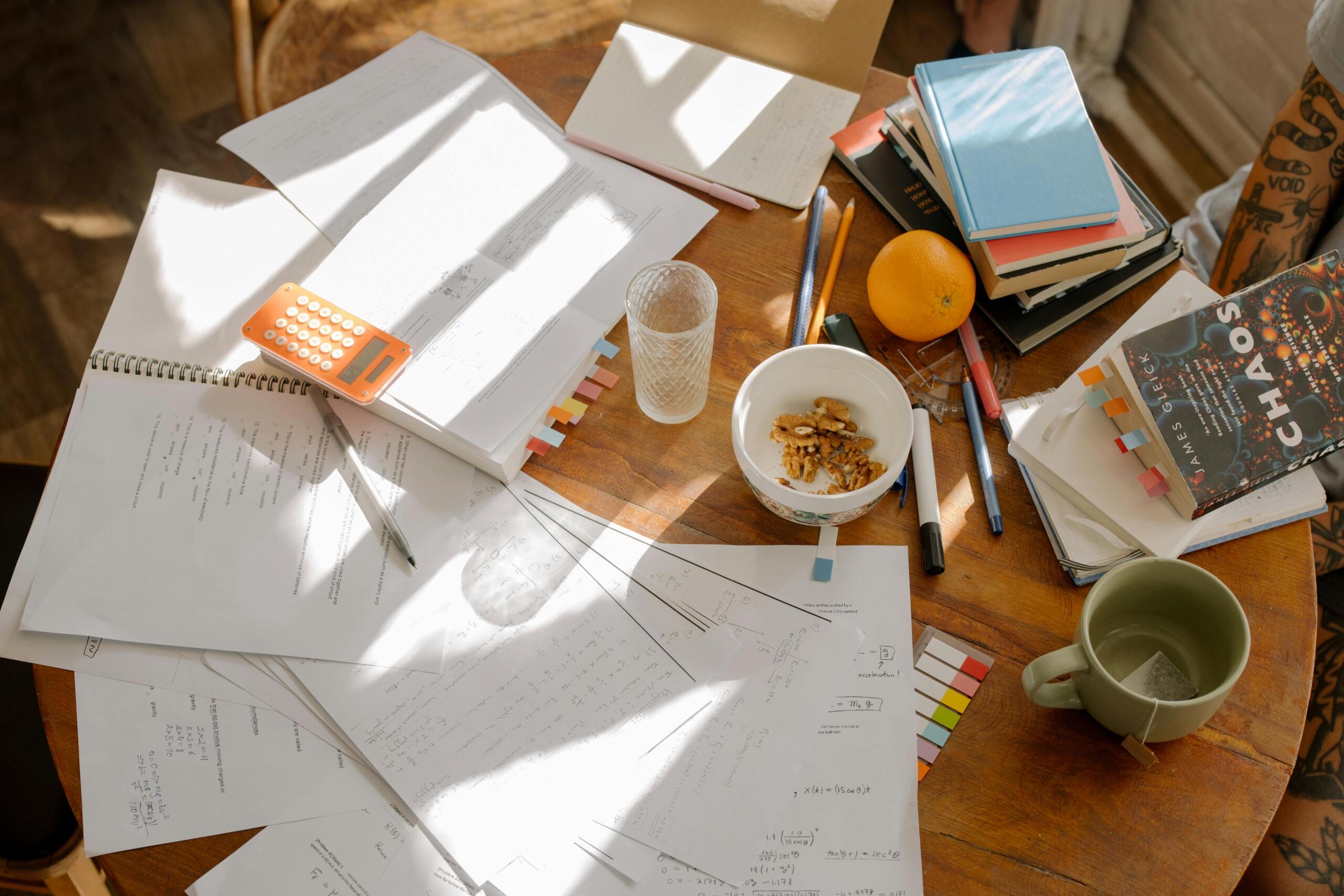 A cluttered study table with books, papers, pens, a calculator, and a bowl of nuts.